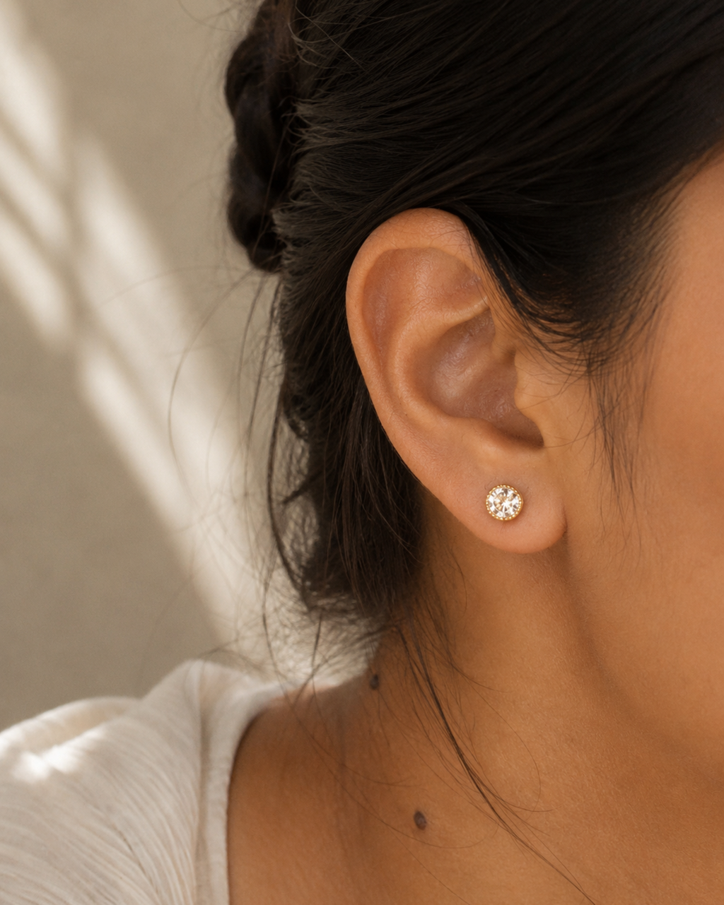 A close-up of a person's ear wearing a small round silver stud earring with a clear faceted gemstone, against a softly lit neutral background.