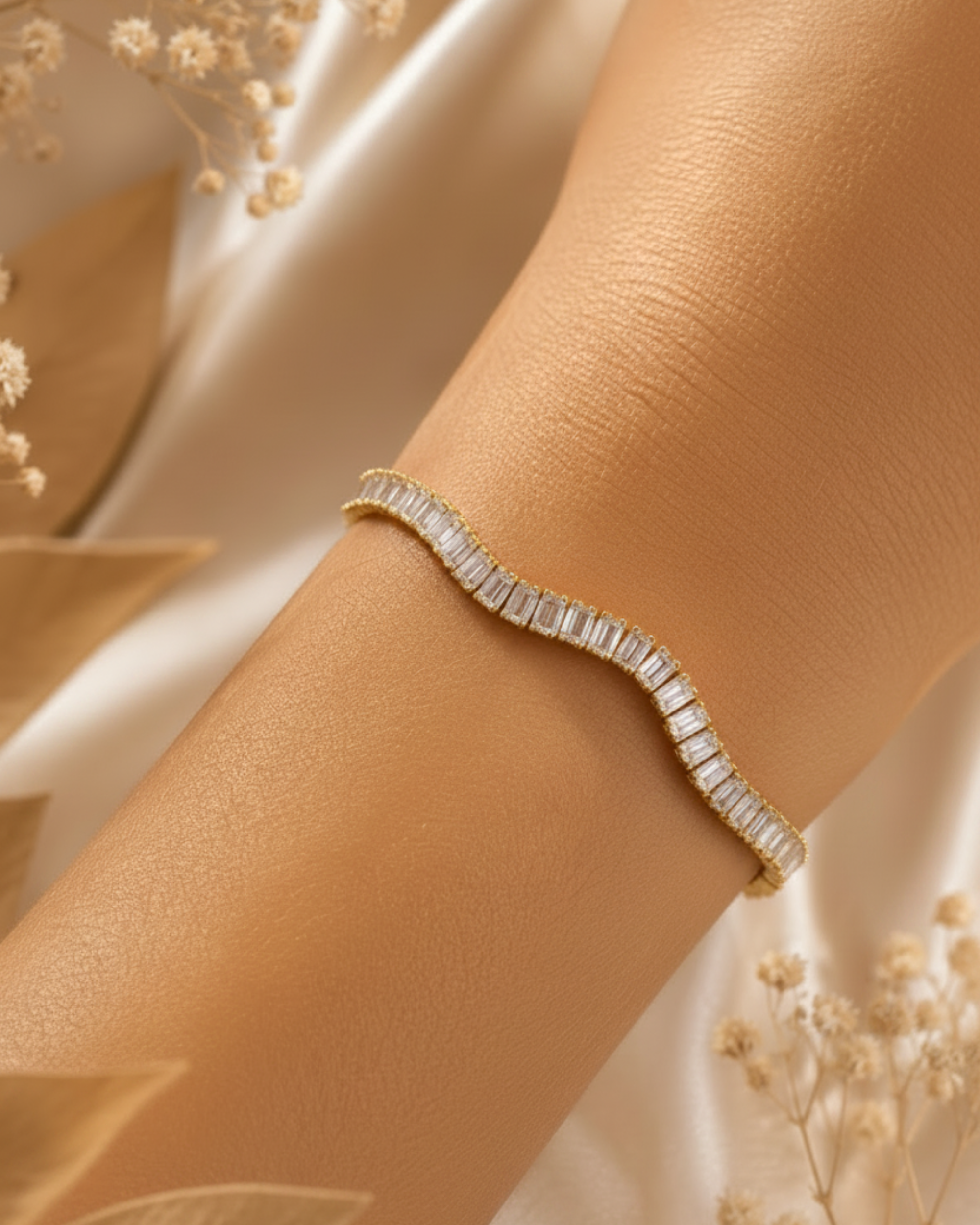 A gold wavy tennis bracelet with baguette-cut clear stones, shown worn on a person's wrist against a soft beige satin background with dried flowers.