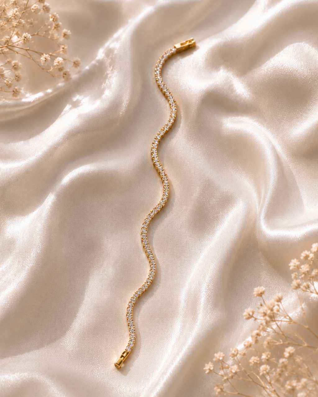 A wavy gold chain bracelet with pavé-set clear stones, styled on draped champagne satin fabric with dried baby's breath flowers.