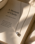 A delicate silver chain necklace with a small clear heart-shaped crystal pendant, draped over the pages of an open book titled 'A Moment of Clarity'.