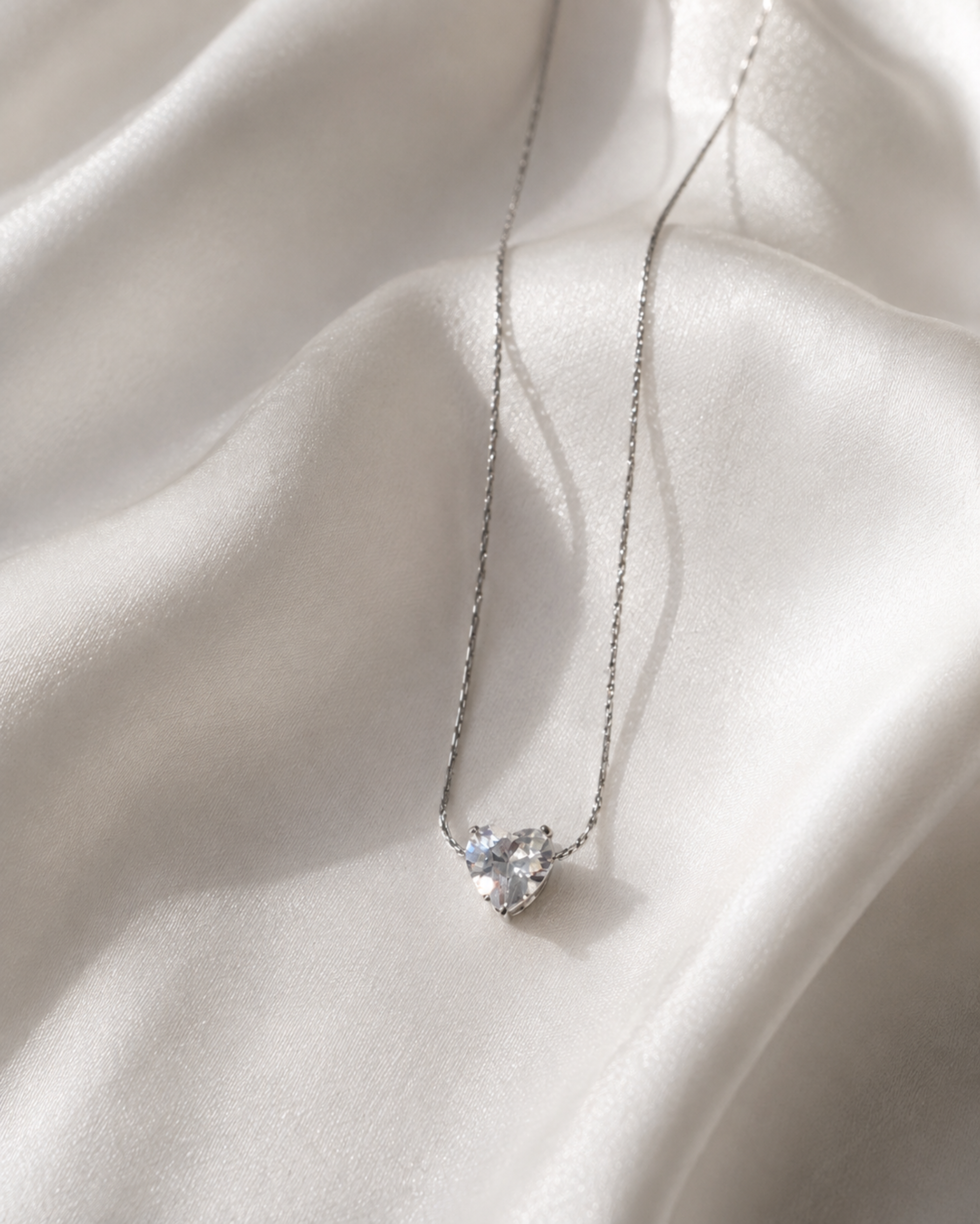 A delicate silver chain necklace with a small heart-shaped clear gemstone pendant, displayed on draped white satin fabric.