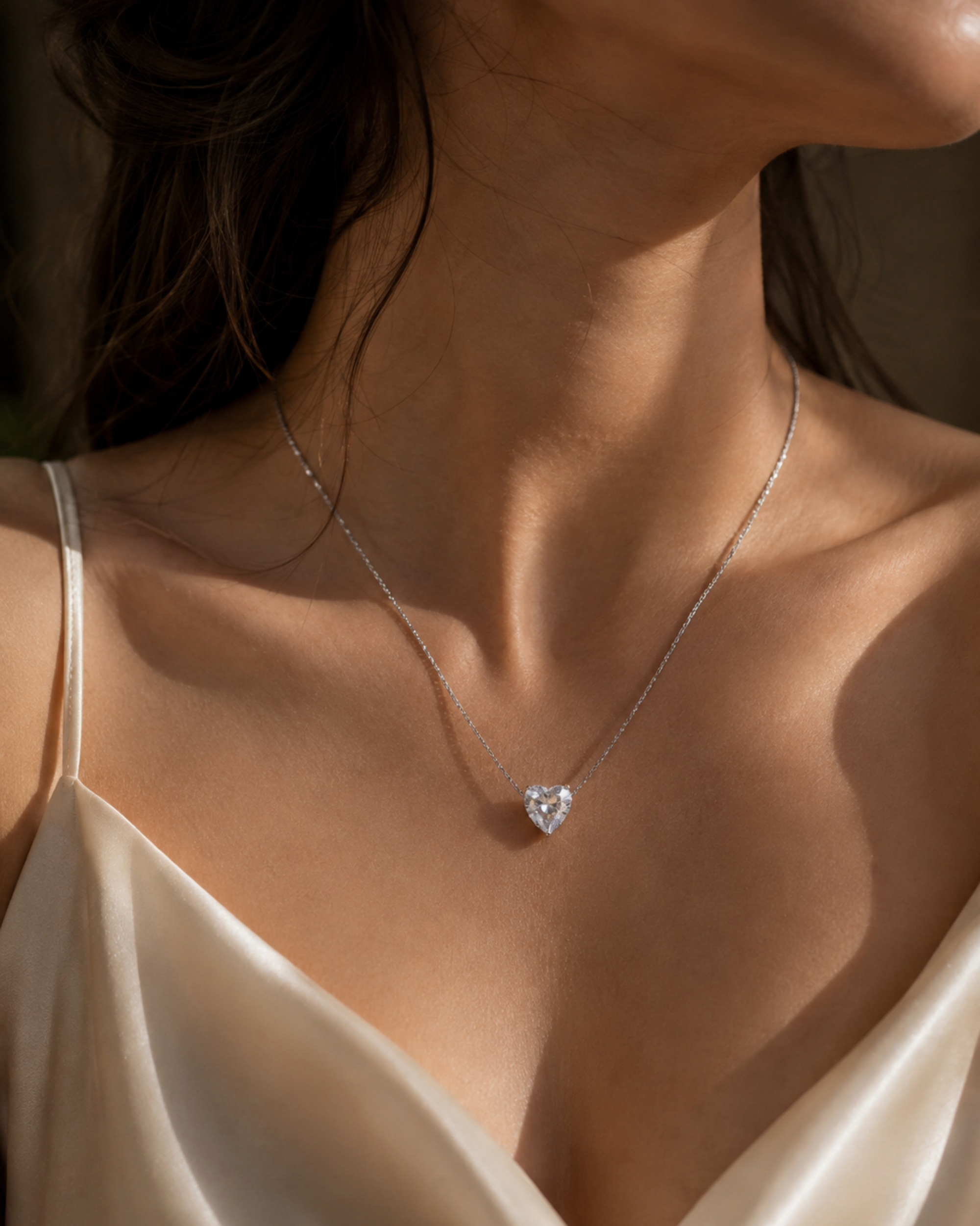 A close-up lifestyle shot of a model wearing a delicate silver chain necklace with a heart-shaped clear gemstone pendant, styled with a cream satin top.