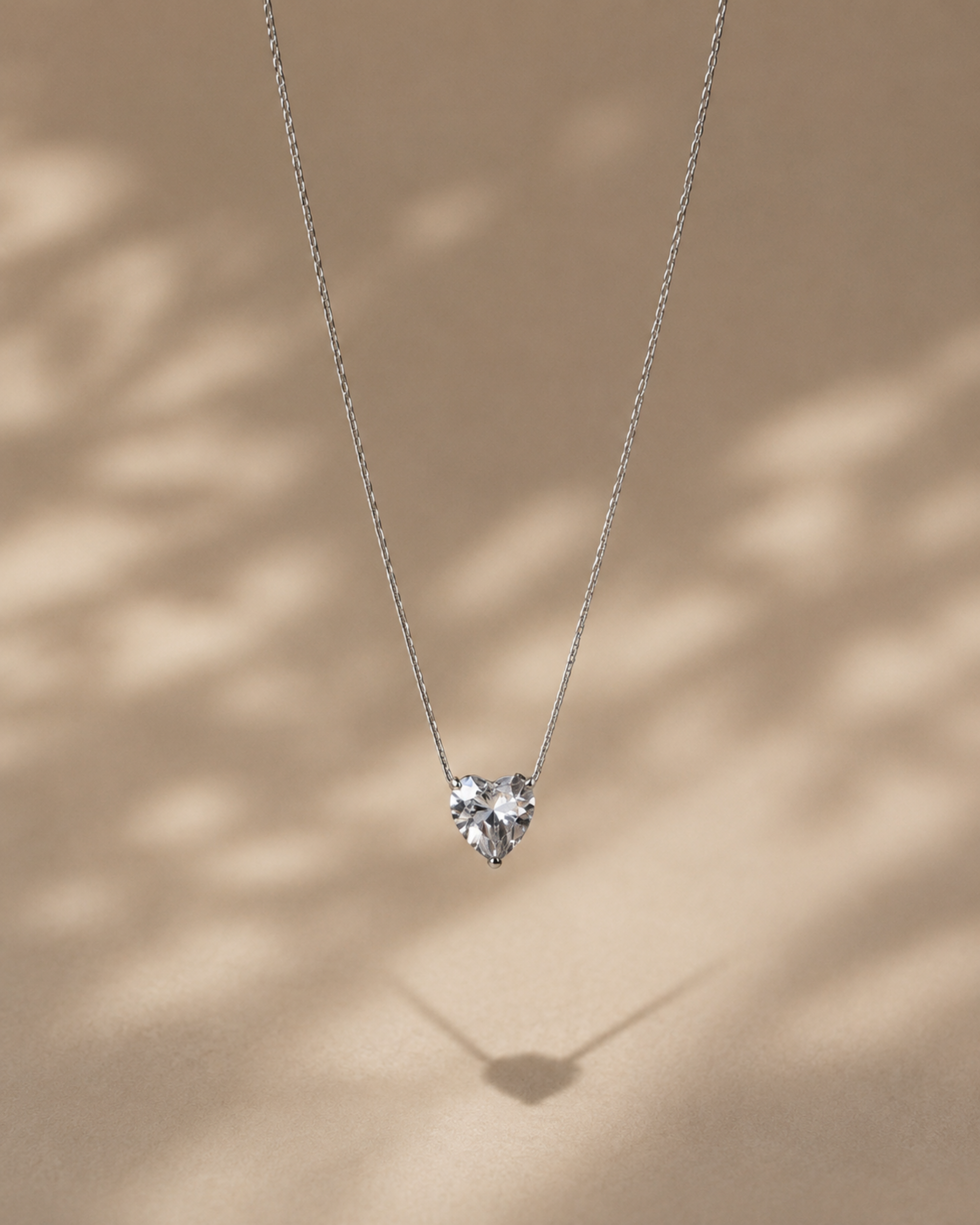 A delicate silver chain necklace with a small heart-shaped clear gemstone pendant, displayed against a warm beige background with soft leaf shadows.