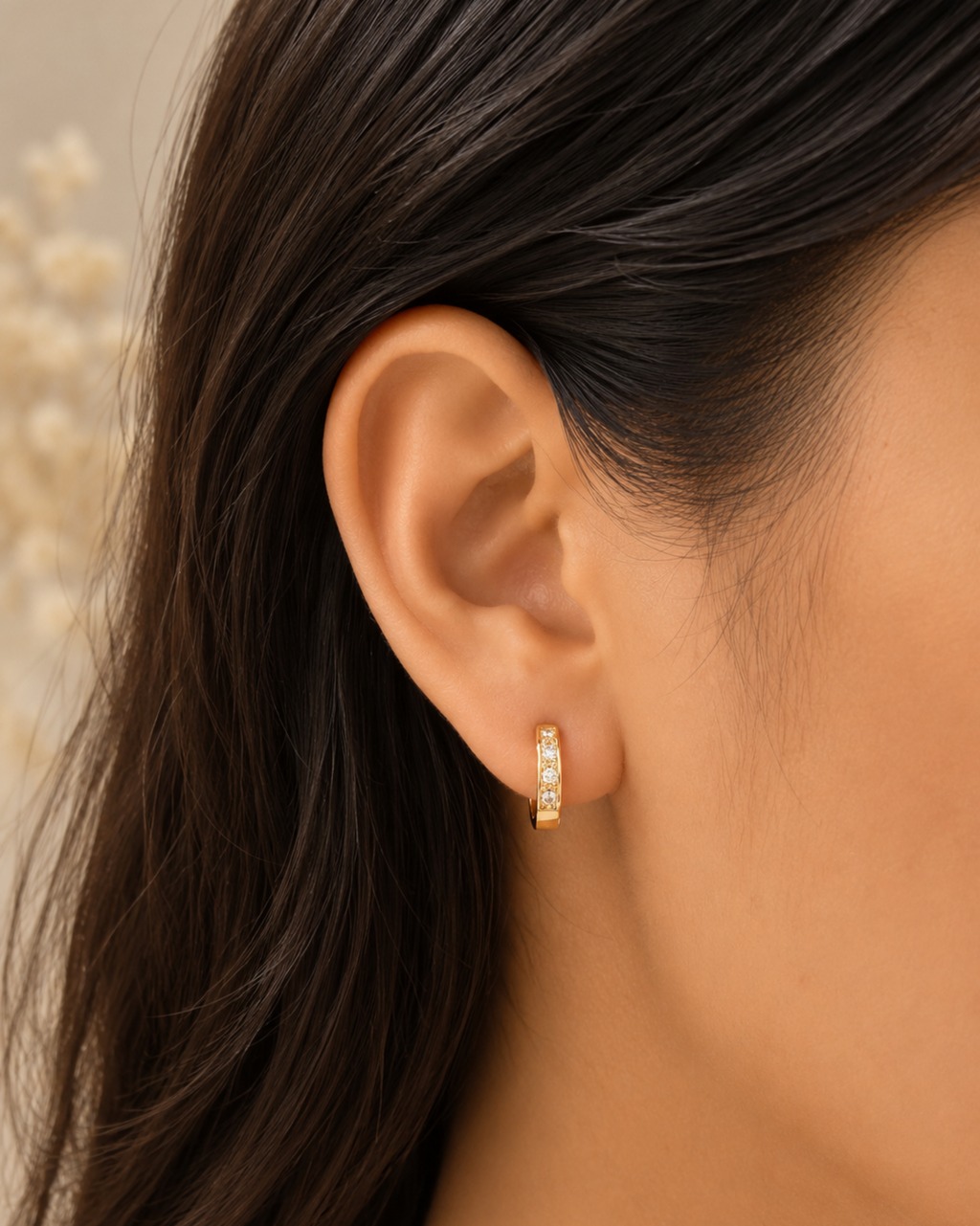 A close-up of a model's ear wearing a small gold huggie hoop earring adorned with clear pavé stones, set against a soft beige background with blurred dried flowers.