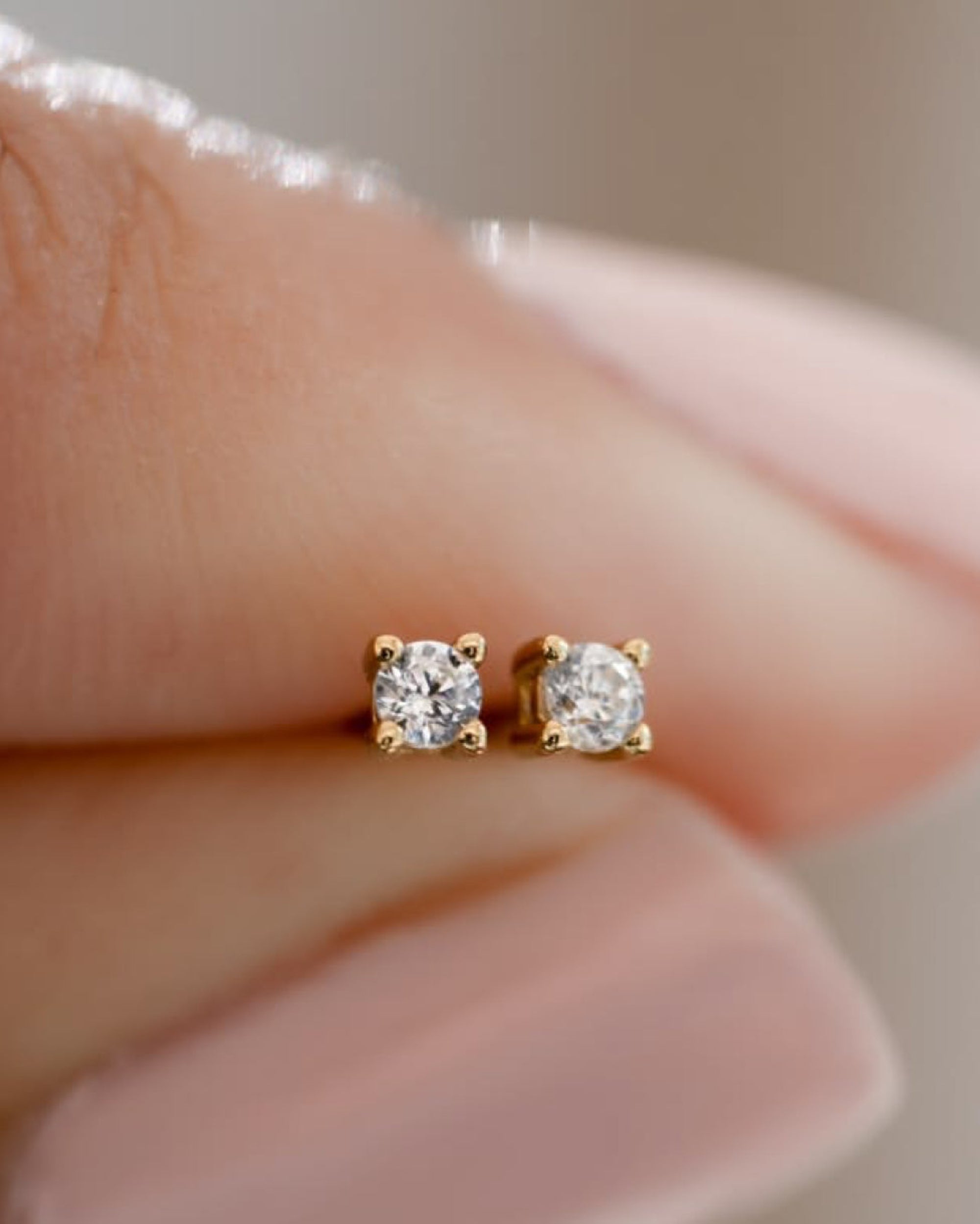 A close-up of a person's fingers holding a pair of small gold stud earrings with clear gemstones.