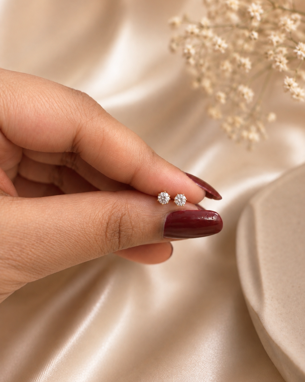 A close-up of a hand with dark burgundy nail polish holding a pair of small silver stud earrings with clear stones, set against a soft beige satin fabric background with dried flowers.