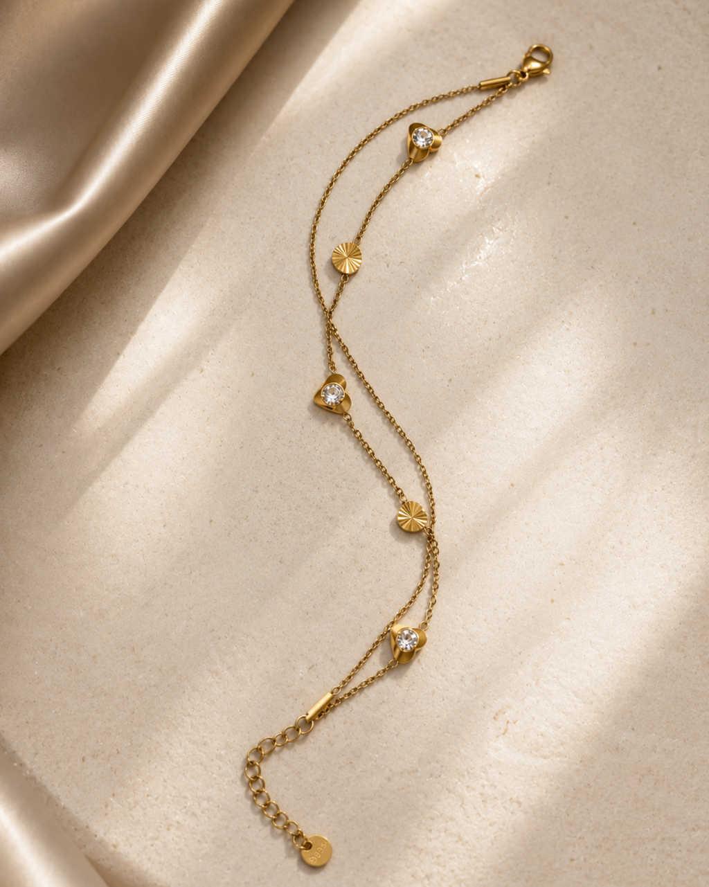 A delicate gold chain anklet with small heart-shaped charms, some adorned with clear stones, displayed on a textured beige surface with soft satin fabric in the background.