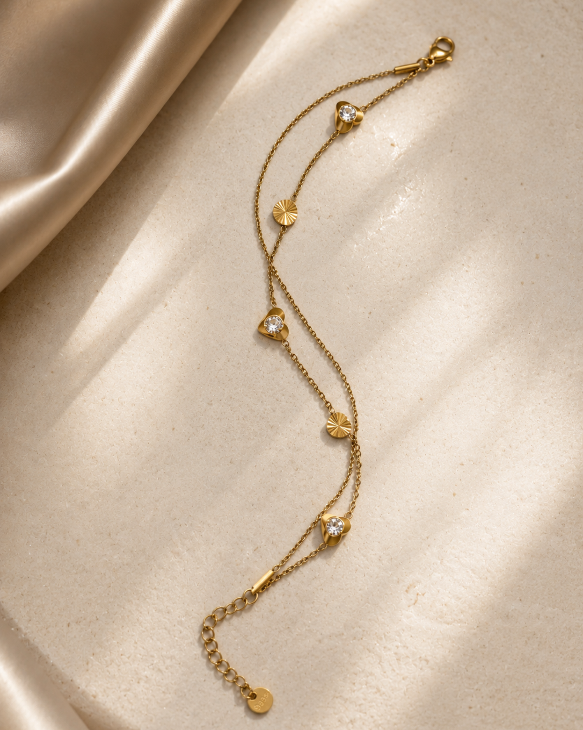 A delicate gold chain anklet with small heart-shaped charms, some adorned with clear stones, displayed on a textured beige surface with soft satin fabric in the background.