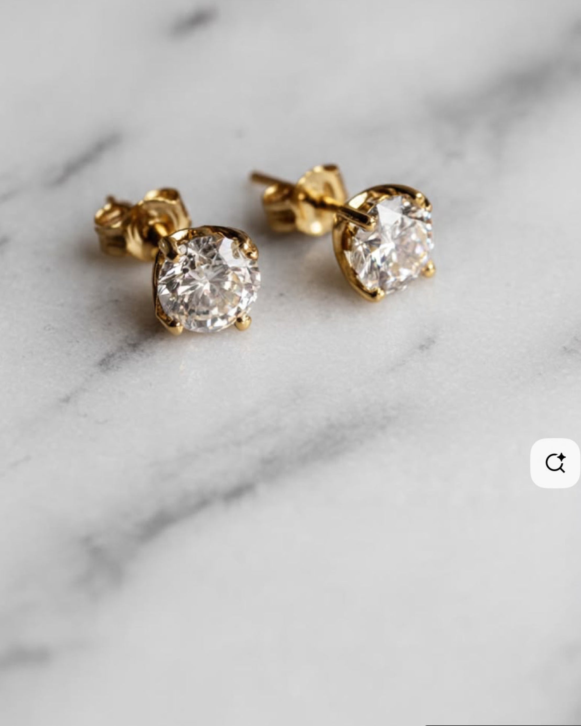 A pair of yellow gold stud earrings with round clear gemstones in a prong setting, resting on a white marble surface.