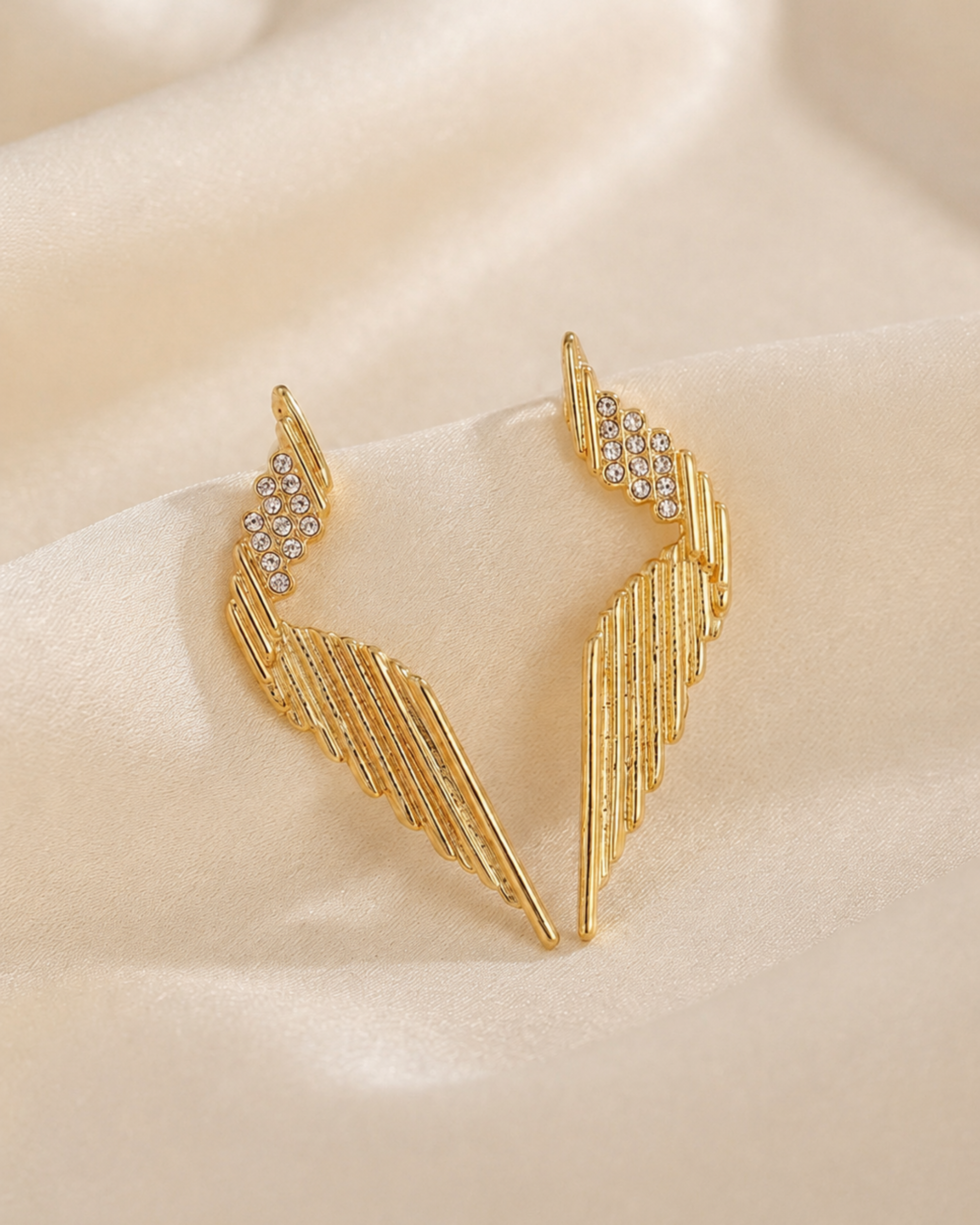 Gold-toned wing-shaped earrings with small clear rhinestone clusters at the top, displayed on a soft beige satin fabric background.