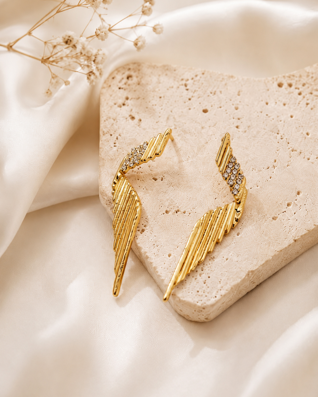 A pair of gold-toned wing-shaped earrings with small clear crystal accents, displayed on a textured beige stone slab with draped cream fabric and dried flowers in the background.