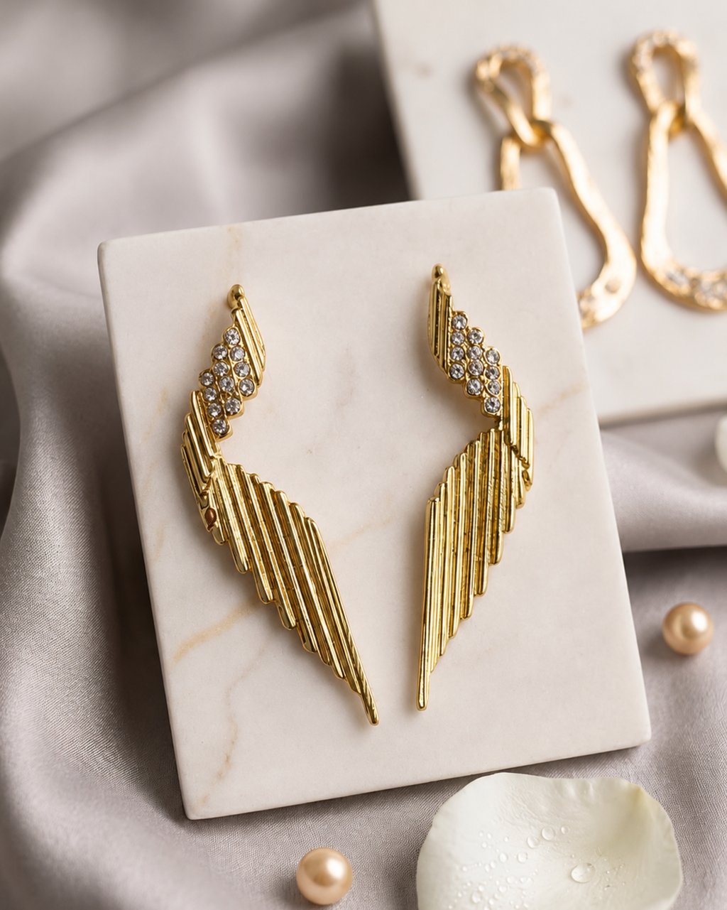 Gold wing-shaped drop earrings with crystal-embellished upper sections, displayed on a white marble card over soft gray fabric with pearl-like beads and a white flower petal.