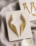 Gold wing-shaped drop earrings with crystal-embellished upper sections, displayed on a white marble card over soft gray fabric with pearl-like beads and a white flower petal.