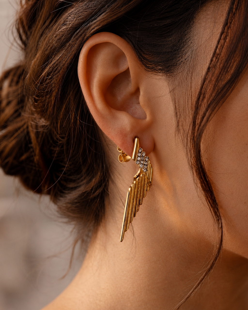 A close-up of a model's ear wearing a gold wing-shaped dangle earring with small clear stones along the top.