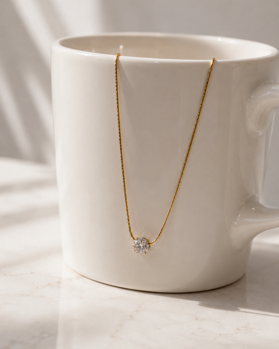 A delicate gold necklace with a small diamond-like flower pendant draped over a white ceramic mug on a light marble surface.