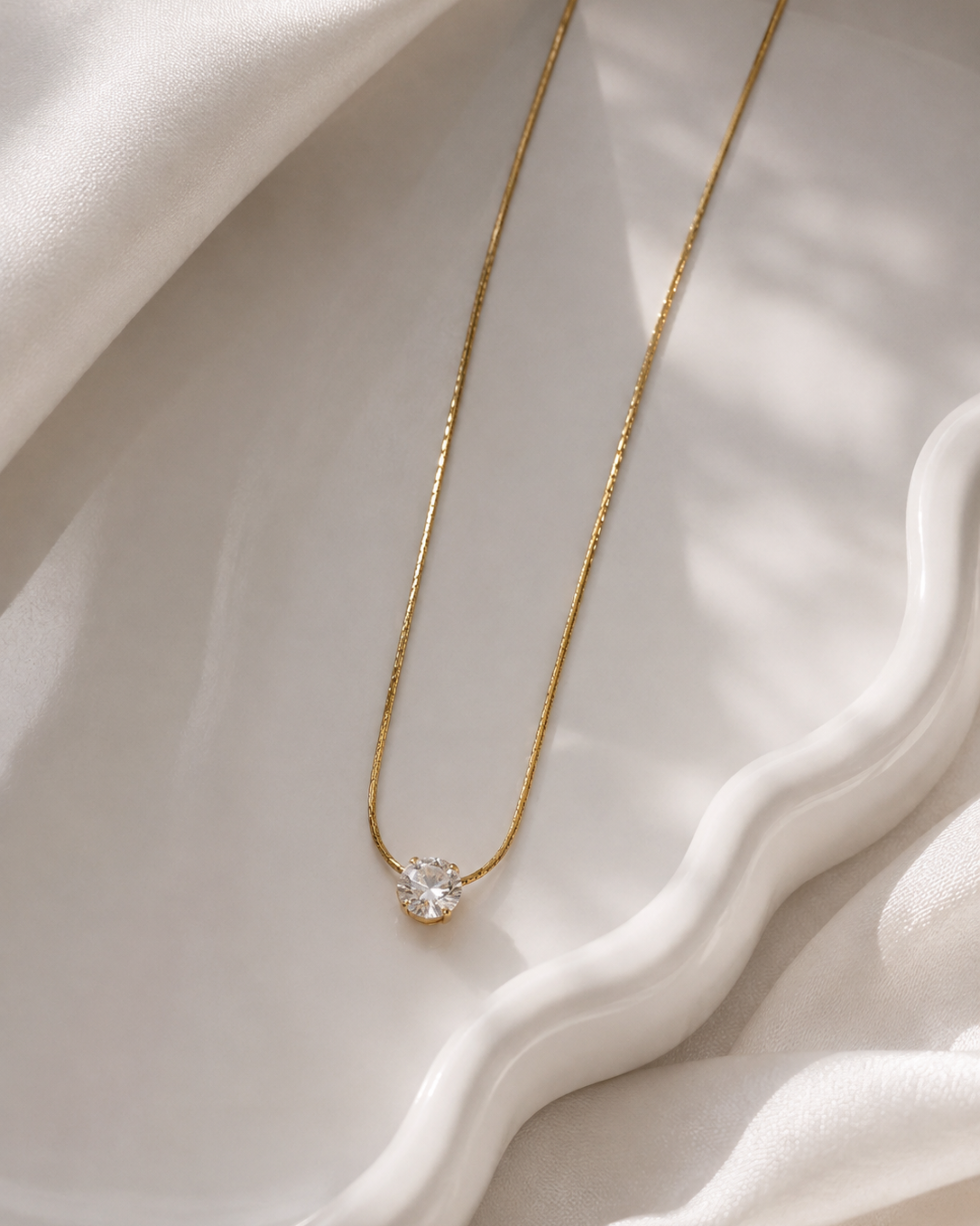 A delicate gold chain necklace with a small gold pendant set with a clear gemstone, displayed on a white ceramic dish with draped white fabric.