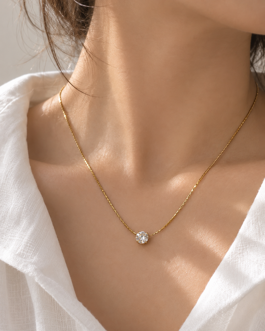 A close-up of a delicate gold chain necklace with a single round clear gemstone pendant, worn on a model's neck over a white blouse.