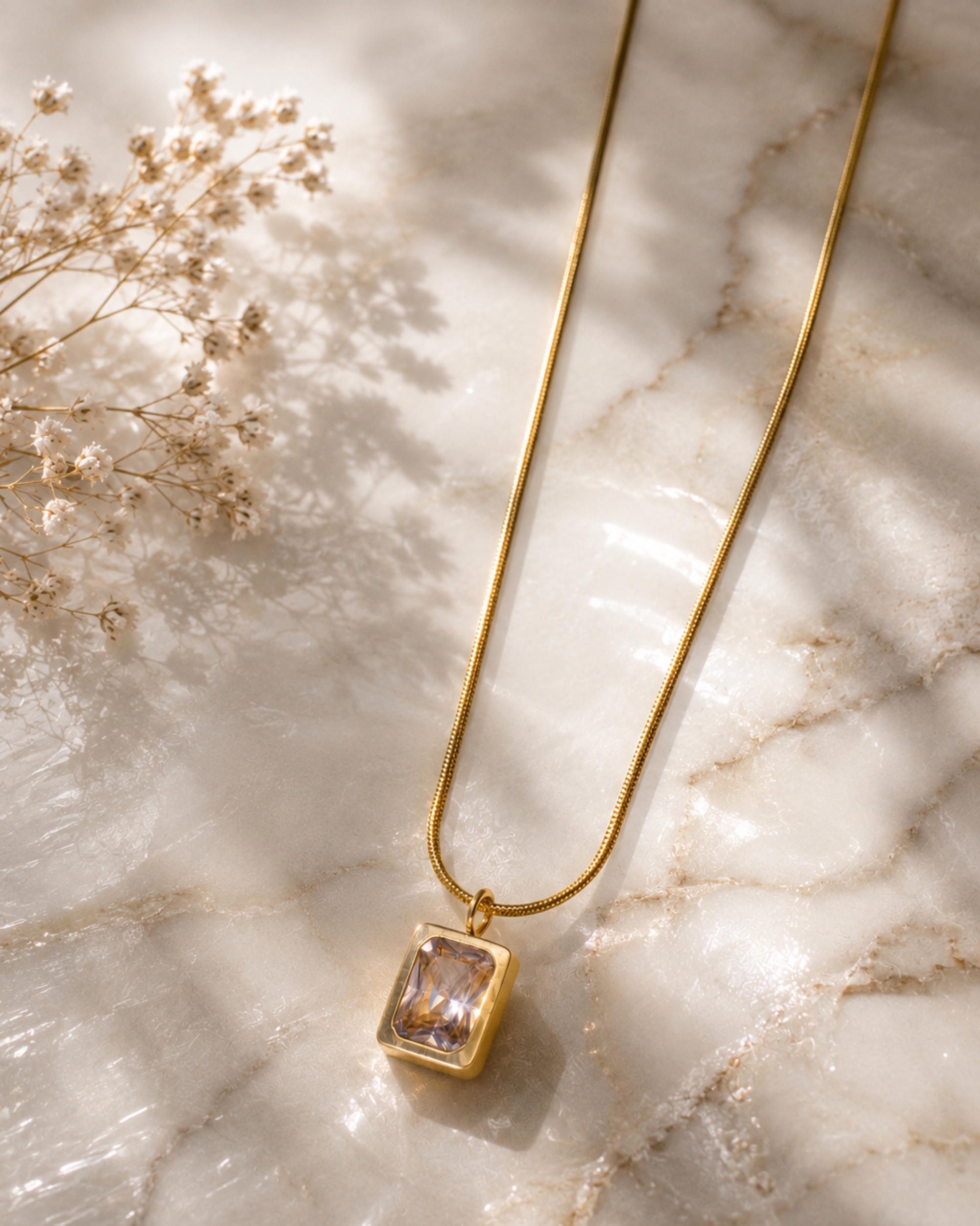 A gold chain necklace with a rectangular gold pendant set with a clear faceted gemstone, displayed on a light beige marble surface with dried baby's breath flowers.