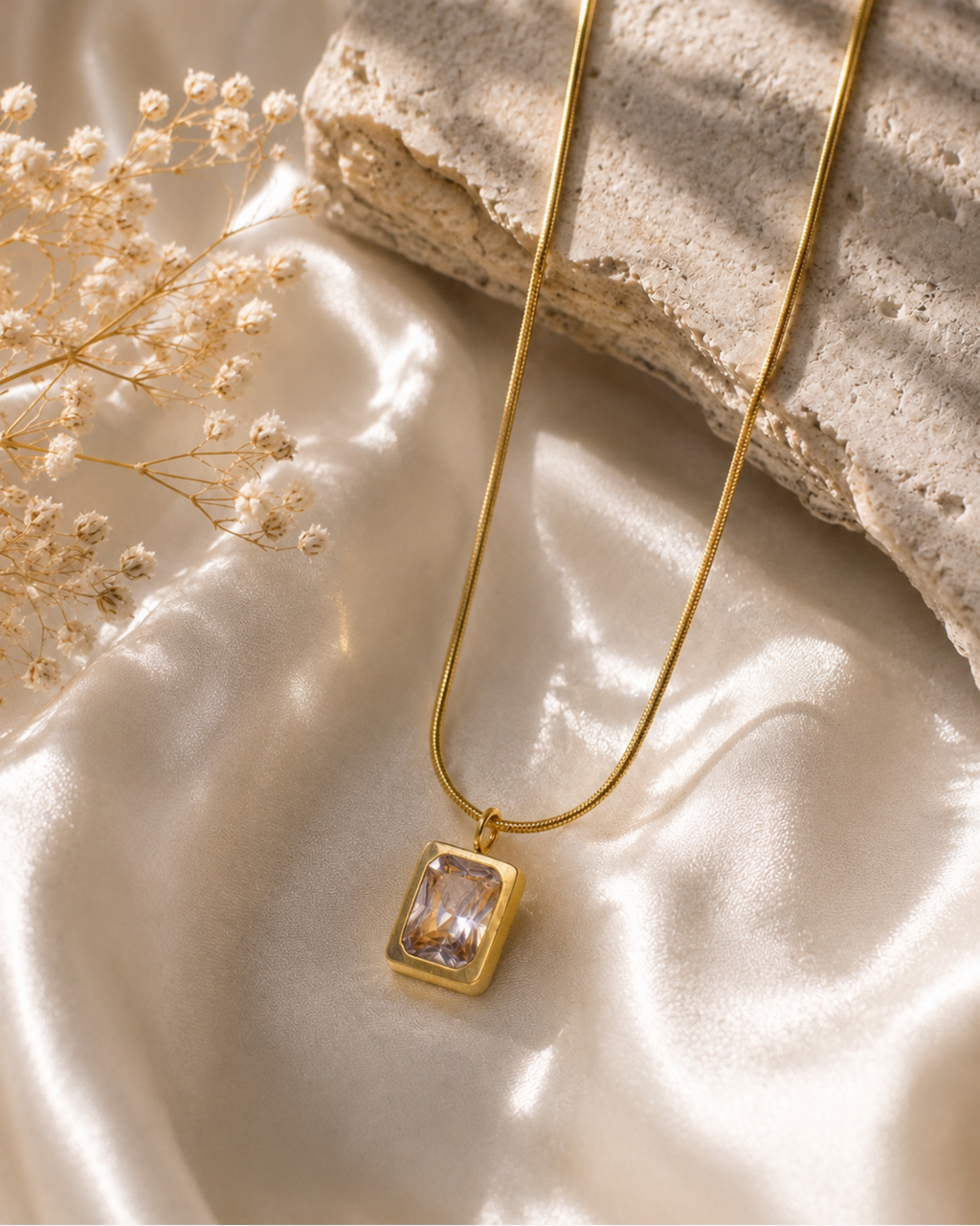A gold pendant necklace with a rectangular clear gemstone set in a gold frame, resting on draped cream satin fabric next to dried baby's breath and a textured stone slab.