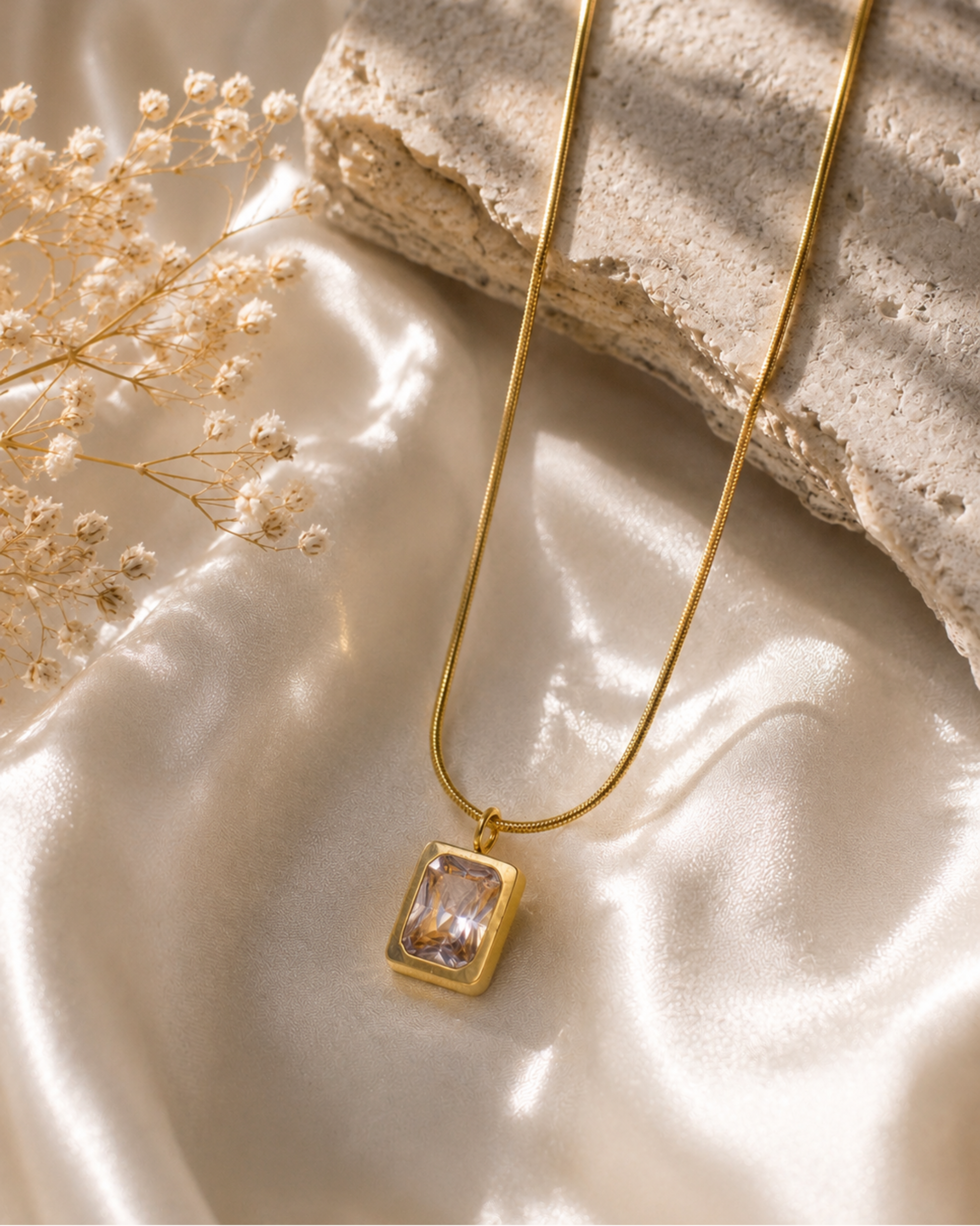A gold pendant necklace with a rectangular clear gemstone set in a gold frame, resting on draped cream satin fabric next to dried baby's breath and a textured stone slab.