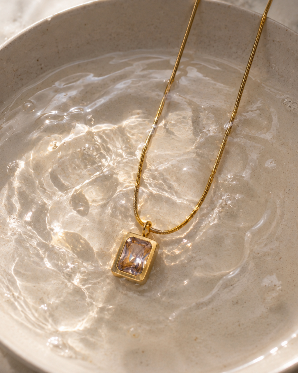A gold chain necklace with a rectangular bezel-set clear gemstone pendant, resting in a shallow dish of water.