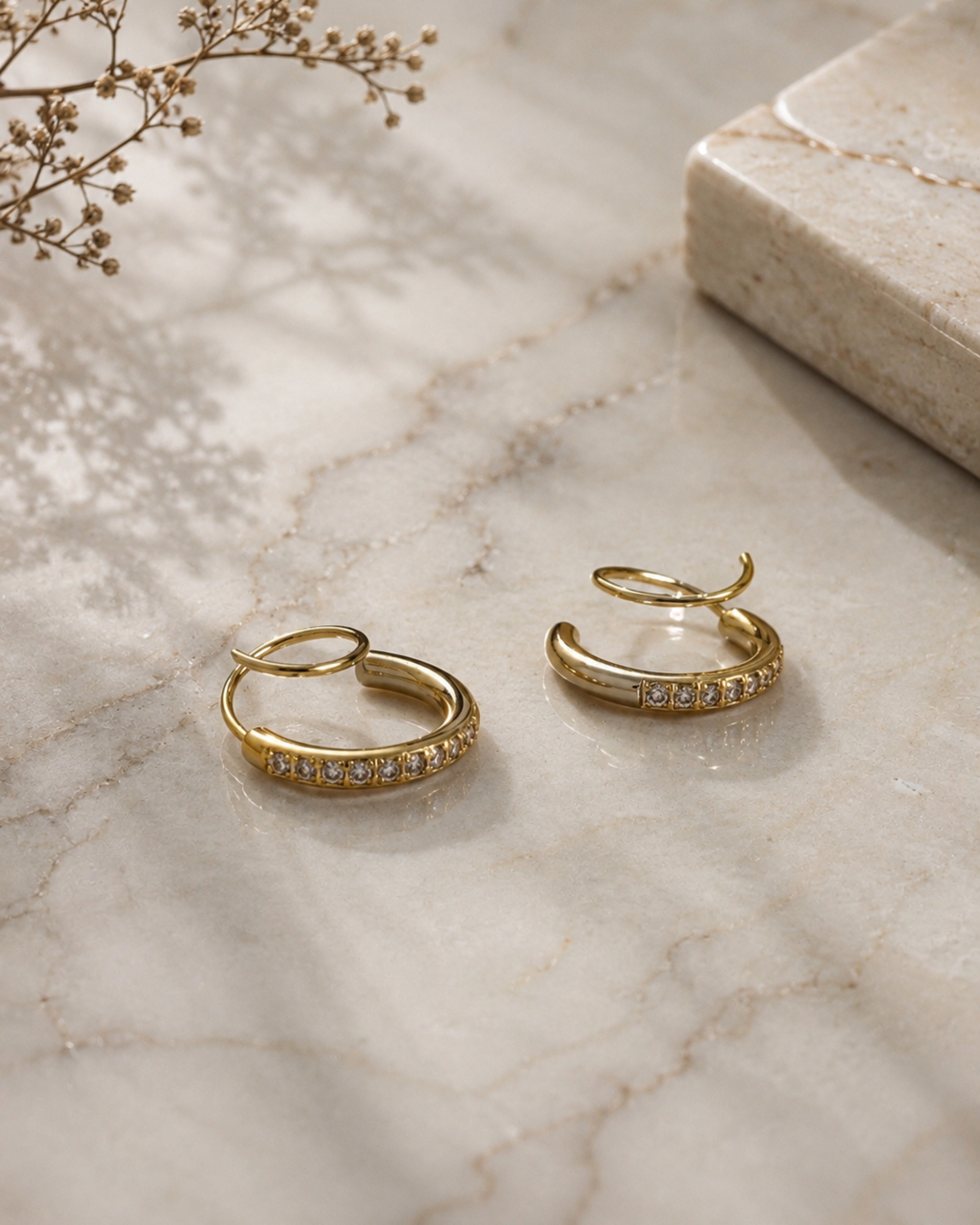 A pair of gold huggie hoop earrings with small clear stones set along the front, displayed on a light marble surface with dried floral stems and a stone slab in the background.