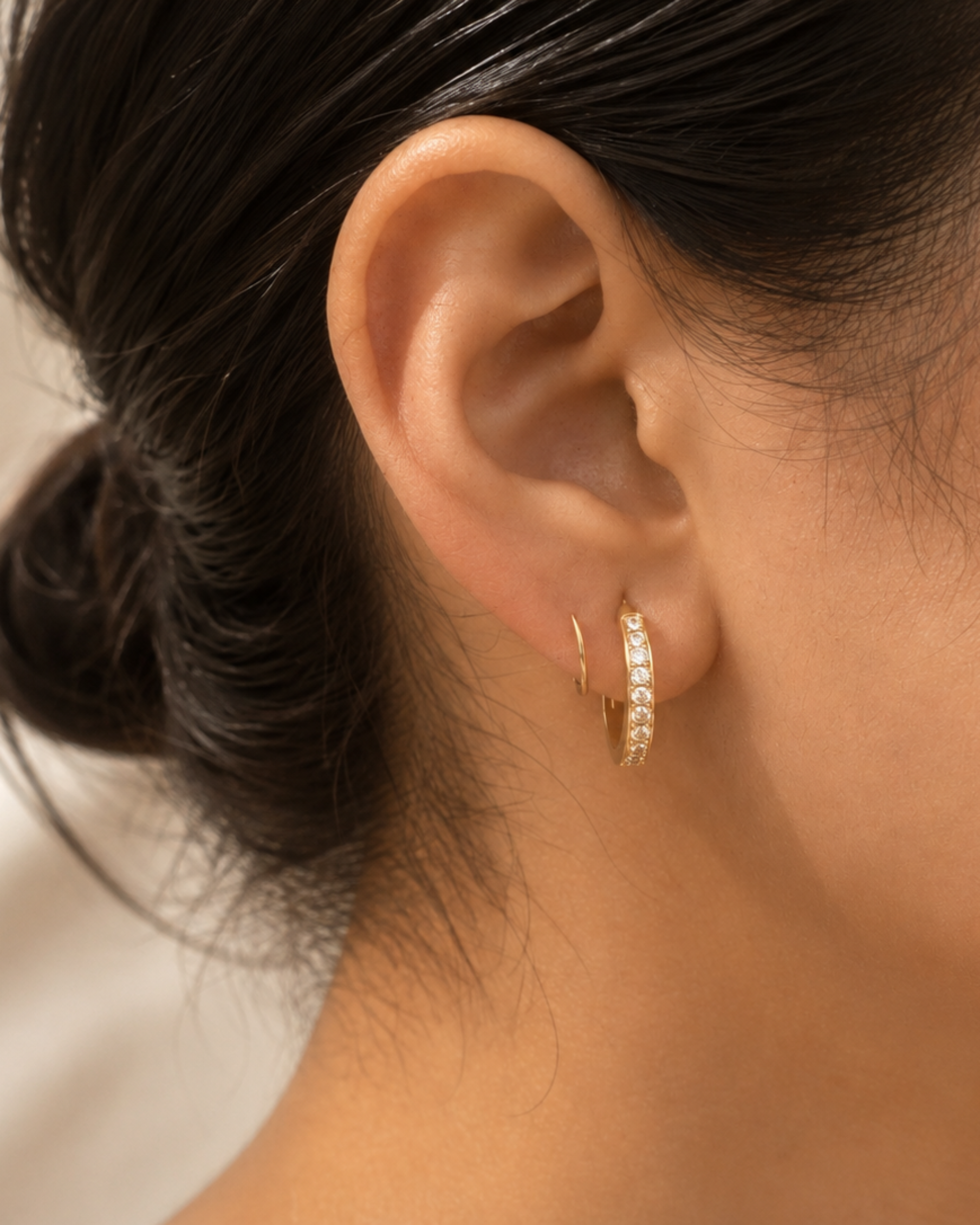 A close-up of a model's ear wearing a small gold huggie hoop earring adorned with clear pavé stones, set against a soft, light beige background.