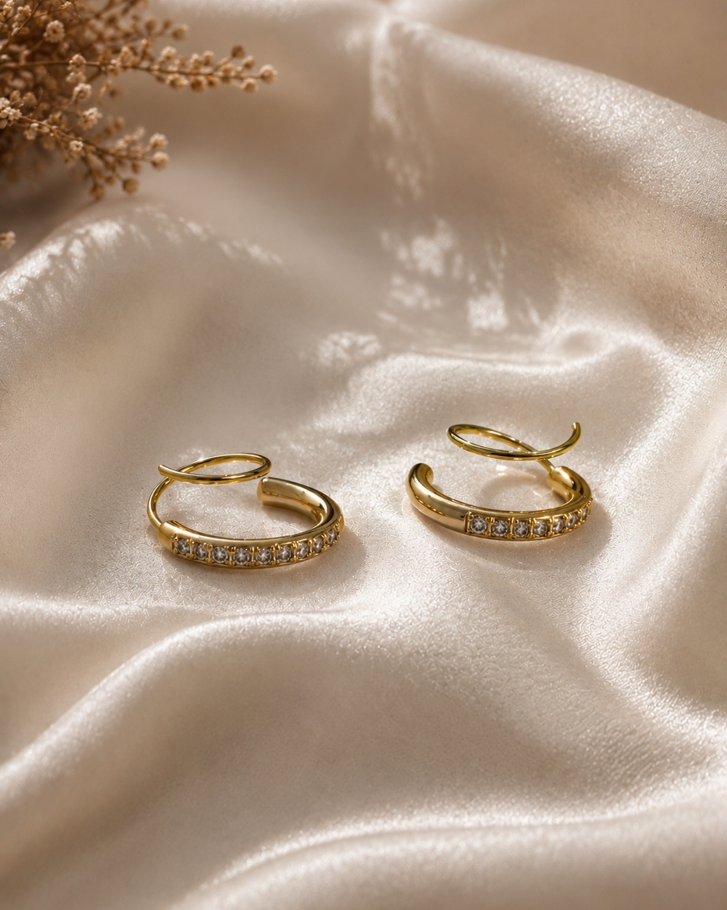 A pair of gold hoop earrings with small clear stones set along the front, displayed on draped champagne satin fabric with dried floral decor in the corner.