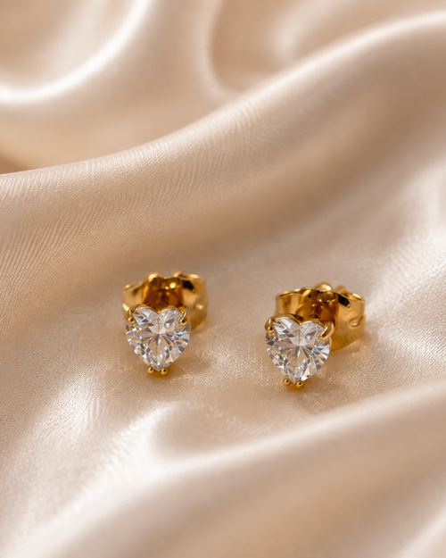 A pair of gold heart-shaped stud earrings with clear heart-shaped gemstones, displayed on a draped beige satin fabric background.