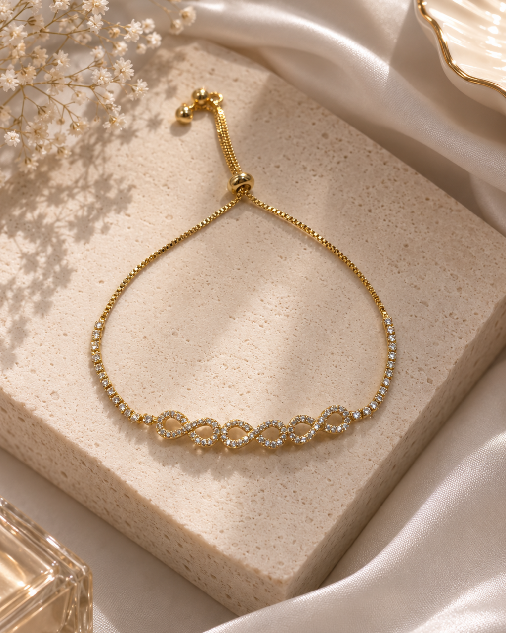 A delicate gold chain bracelet with five interlocking oval links set with clear stones, displayed on a textured beige stone block with white satin fabric and dried flowers.