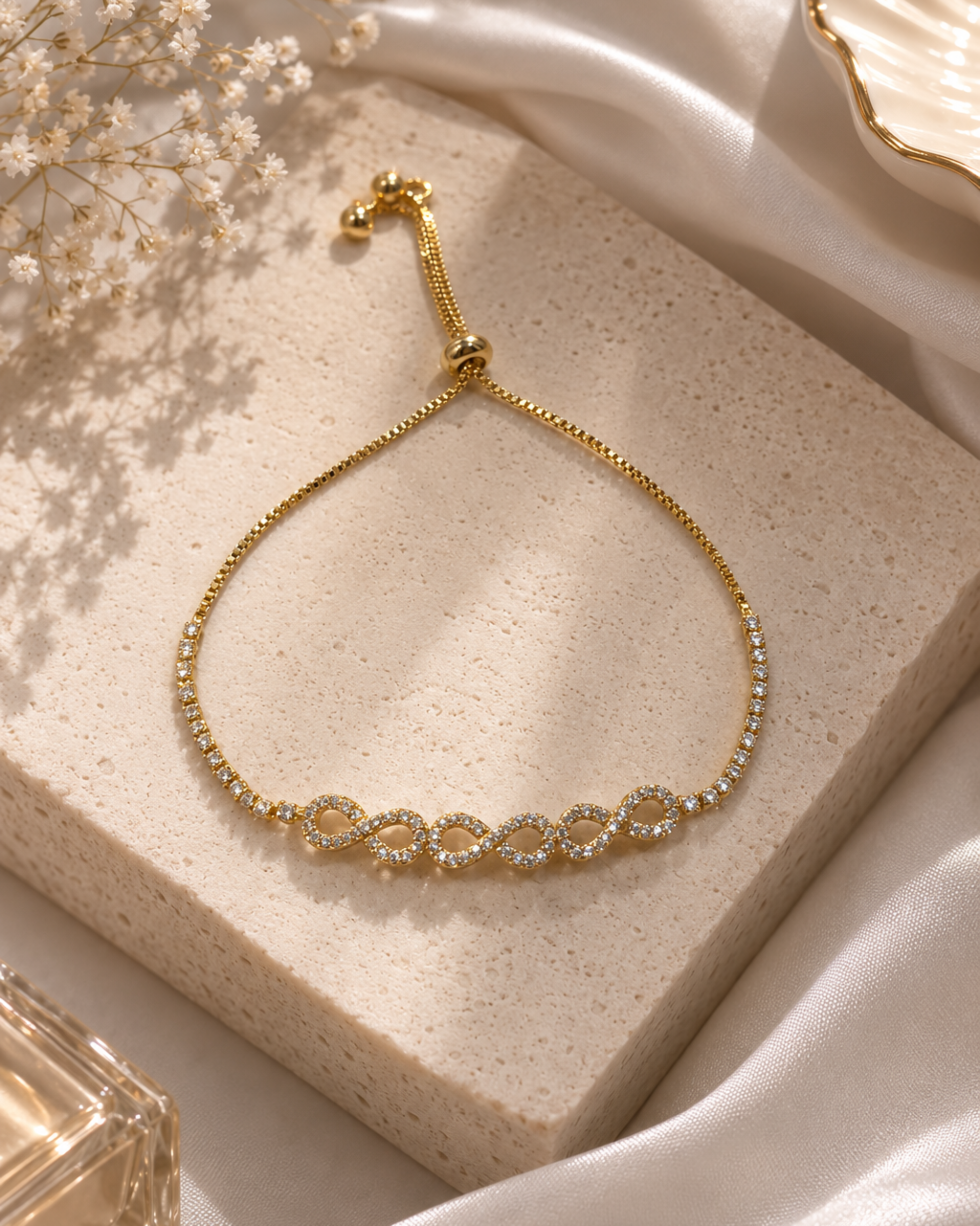 A delicate gold chain bracelet with five interlocking oval links set with clear stones, displayed on a textured beige stone block with white satin fabric and dried flowers.