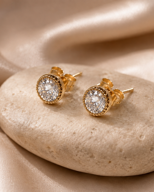 A pair of gold stud earrings with round clear gemstones, displayed on a smooth beige stone over draped satin fabric.