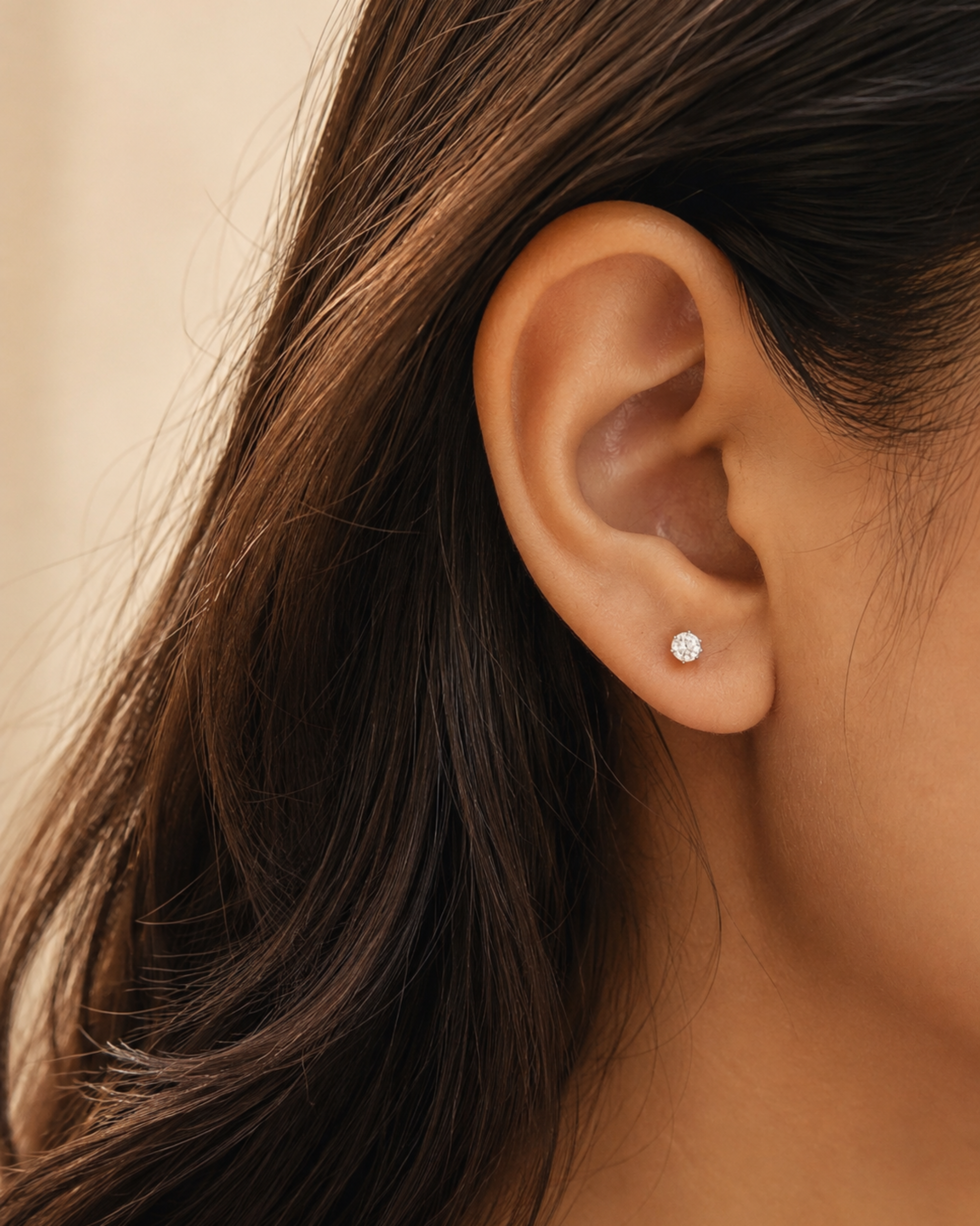 A close-up of a person's ear wearing a small, round, clear stud earring. The earring is set in a silver-toned metal and is shown against the model's warm skin and dark brown hair.
