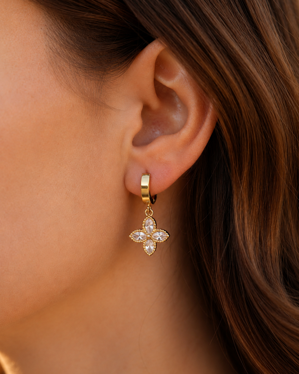 A close-up of a model's ear wearing a gold huggie hoop earring with a dangling four-petal flower charm set with clear rhinestones.