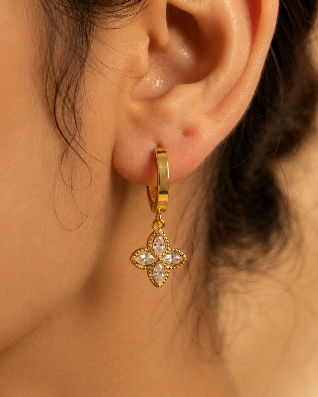 A close-up of a gold huggie hoop earring with a dangling clover-shaped charm set with clear crystals, shown worn on a person's ear.