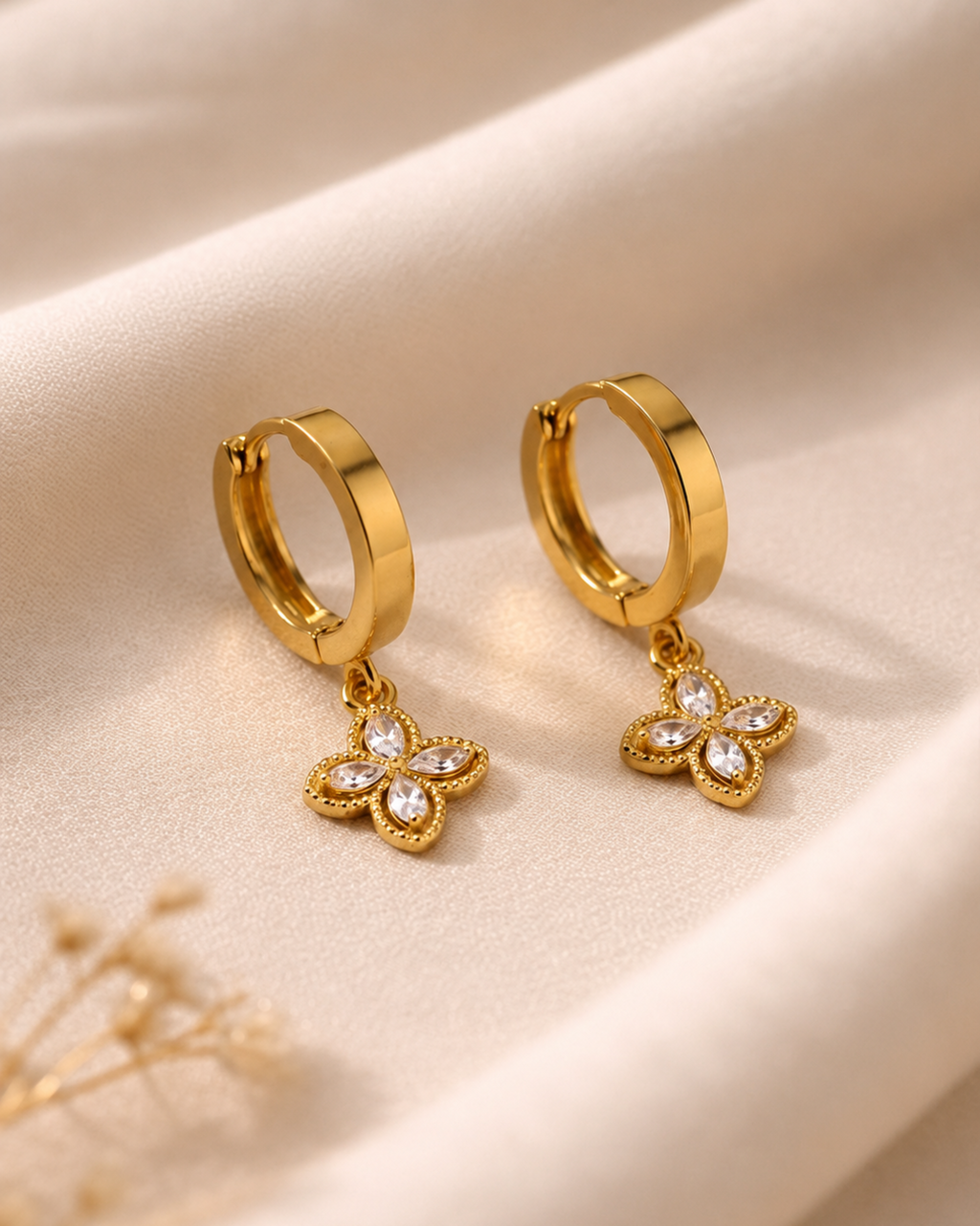 A pair of gold huggie hoop earrings with dangling four-butterfly flower charms set with clear crystals, displayed on a draped beige fabric background.