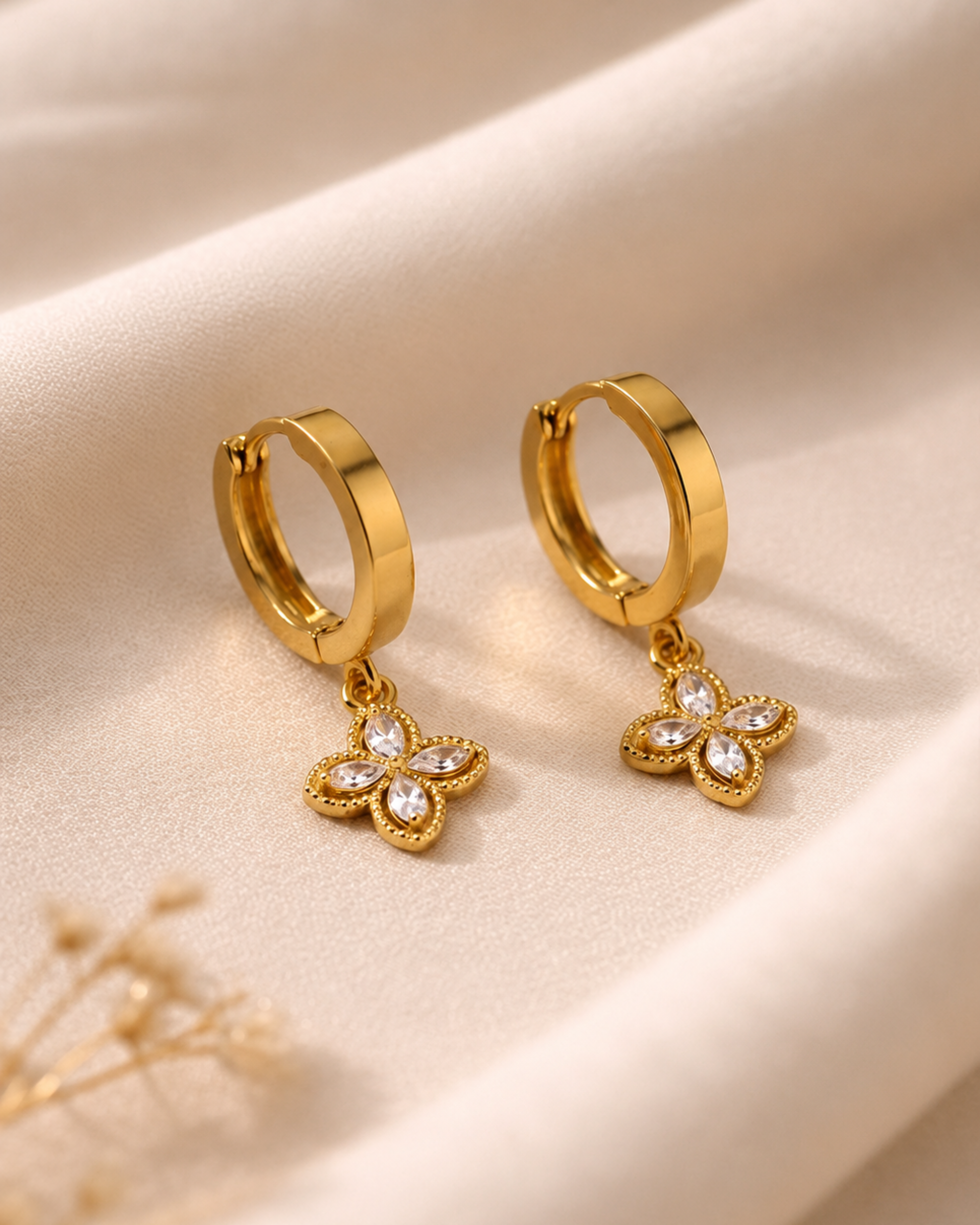 A pair of gold huggie hoop earrings with dangling four-butterfly flower charms set with clear crystals, displayed on a draped beige fabric background.