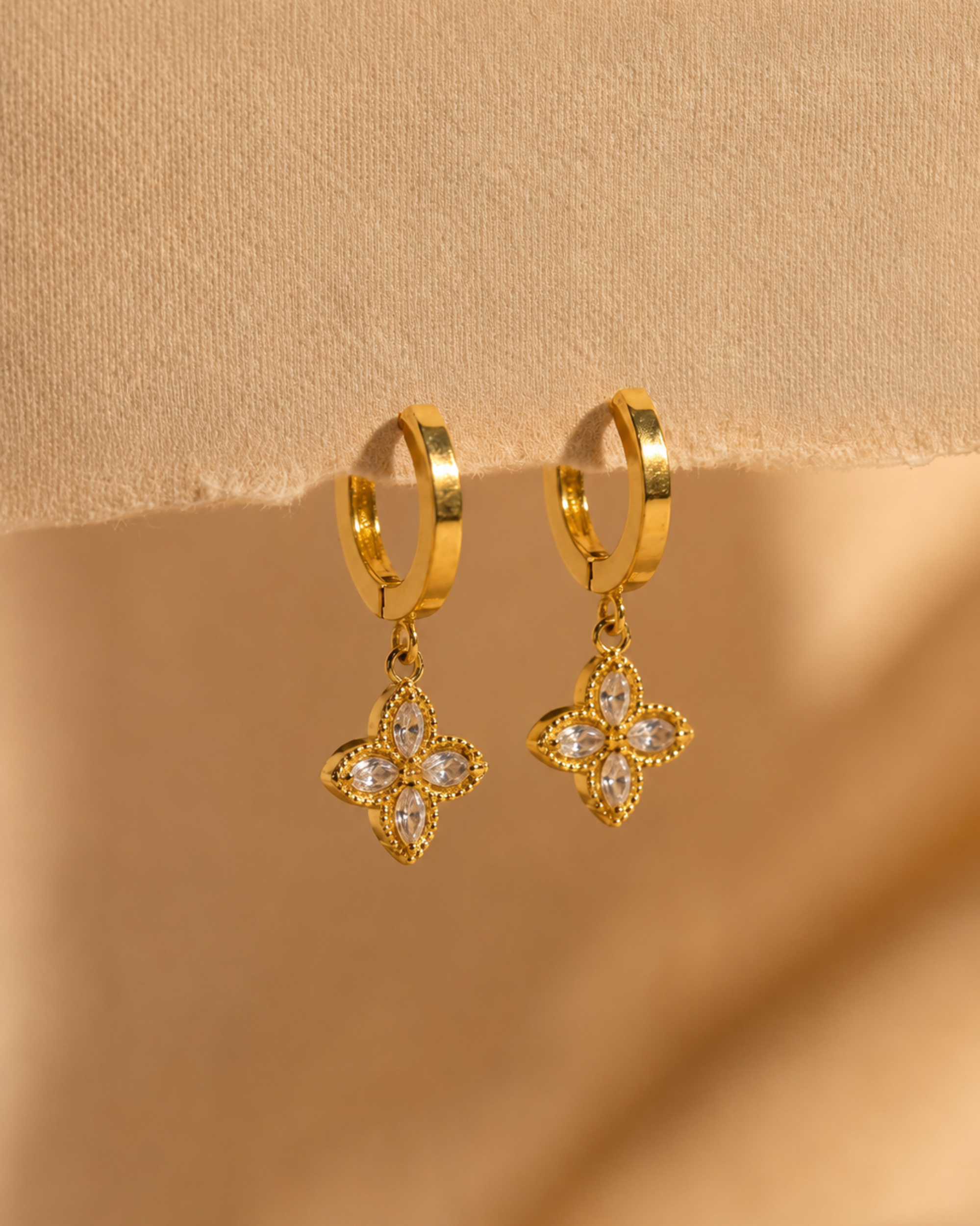 A pair of gold huggie hoop earrings with dangling four-leaf clover charms set with clear crystals, displayed on a textured beige fabric background.