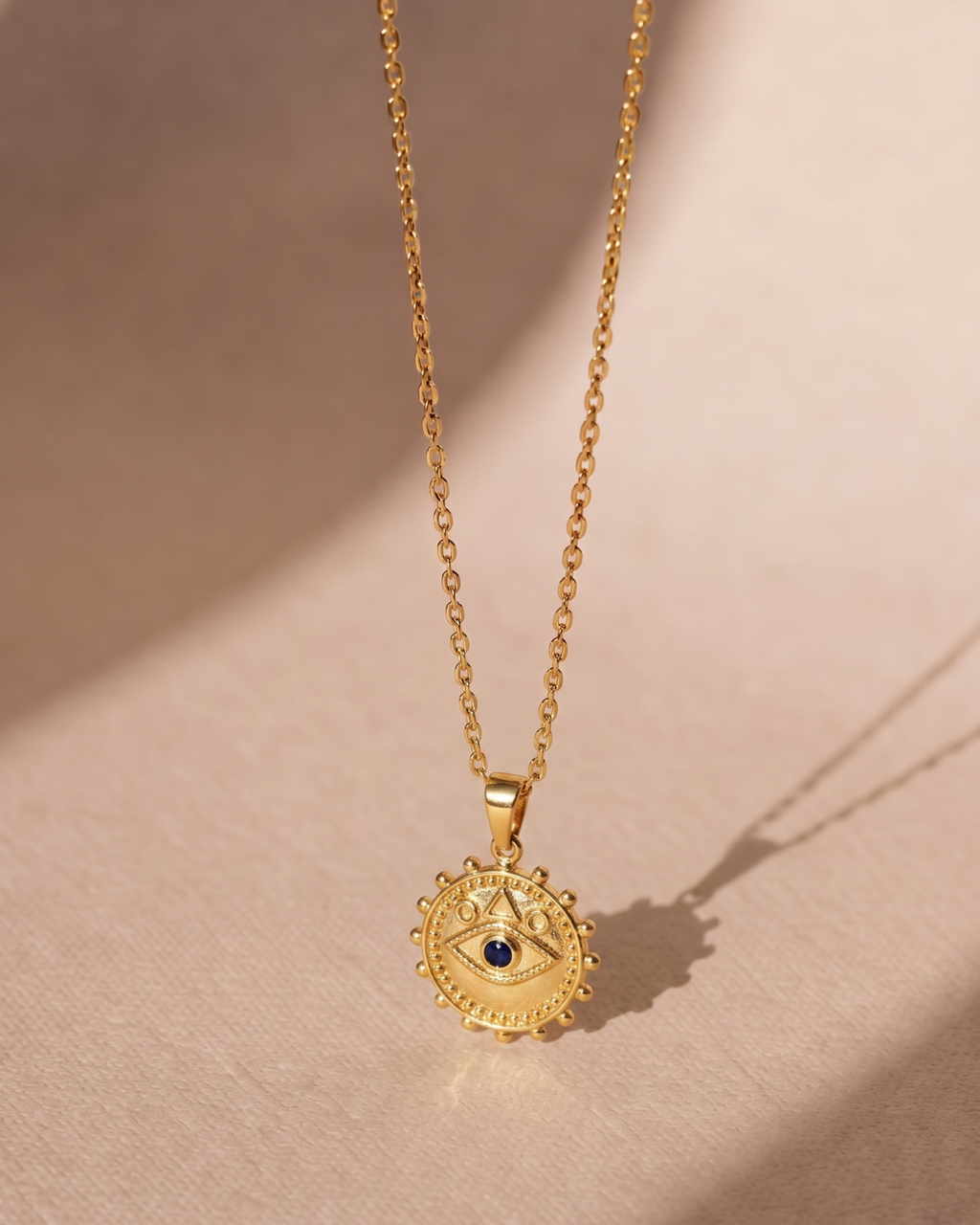 A gold-toned chain necklace with a circular pendant featuring an eye motif and a small dark blue stone at the center, displayed on a warm beige surface with soft shadows.