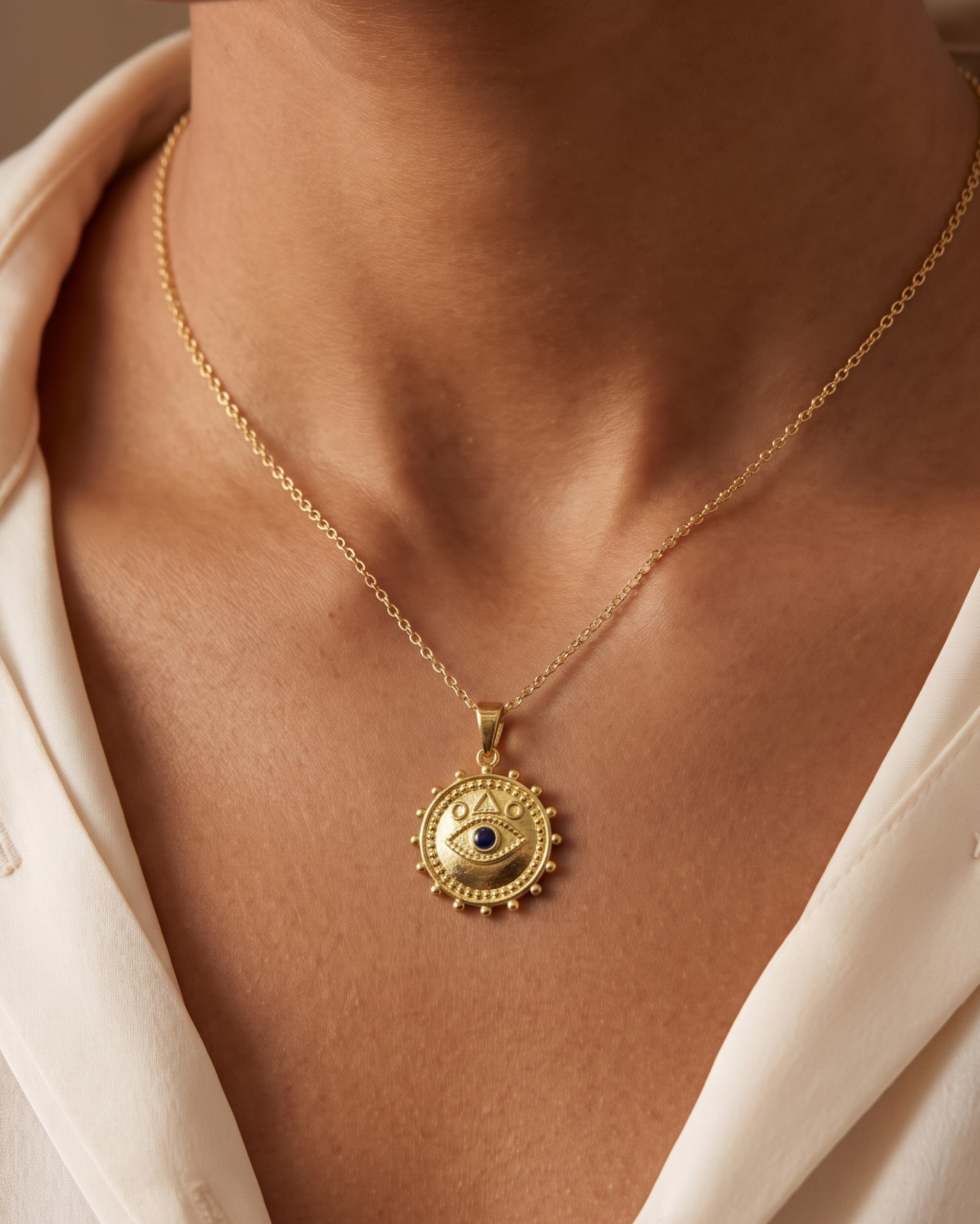 A close-up of a model wearing a delicate gold chain necklace with a circular sun-and-eye pendant, styled with a cream-colored collared shirt.