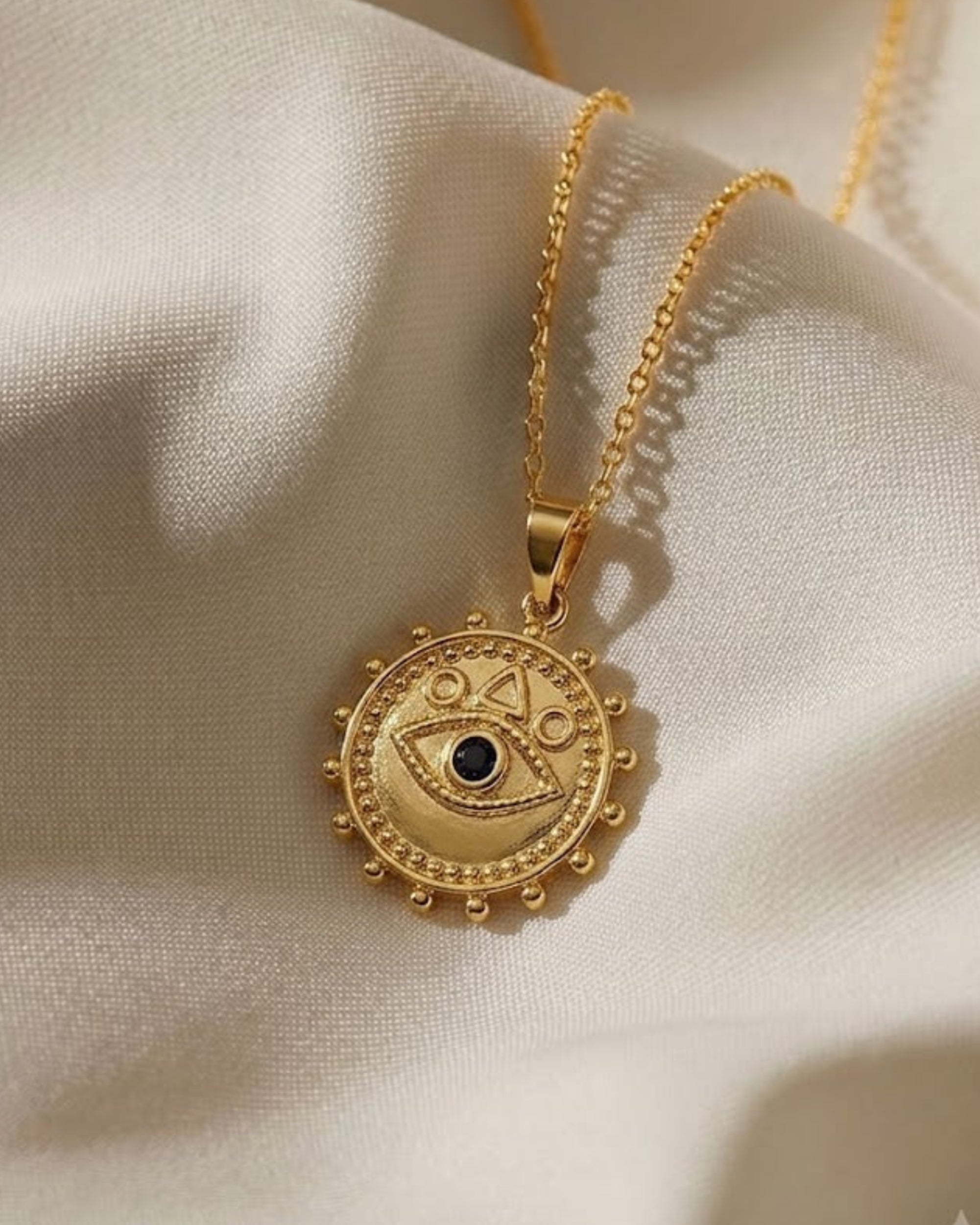 A gold pendant necklace featuring an eye of protection design with a small black stone in the center, resting on draped cream-colored satin fabric.