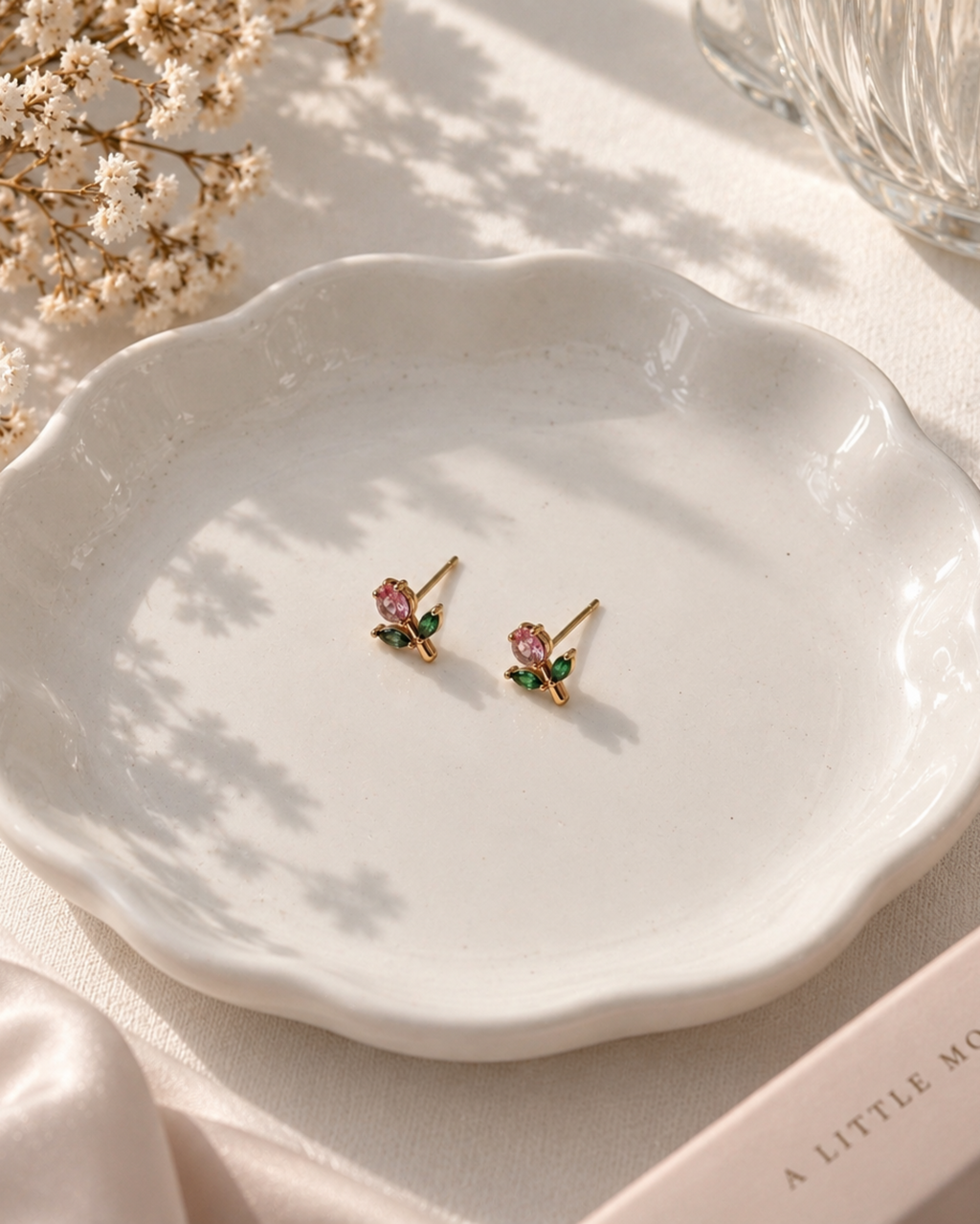 A pair of gold rose stud earrings with pink and green gemstones, displayed on a white scalloped dish with dried flowers and soft sunlight.