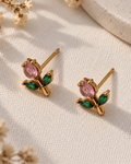 A pair of gold butterfly stud earrings with marquise-cut pink stones and green leaf accents, displayed on a textured cream fabric with dried flowers and soft shadows.
