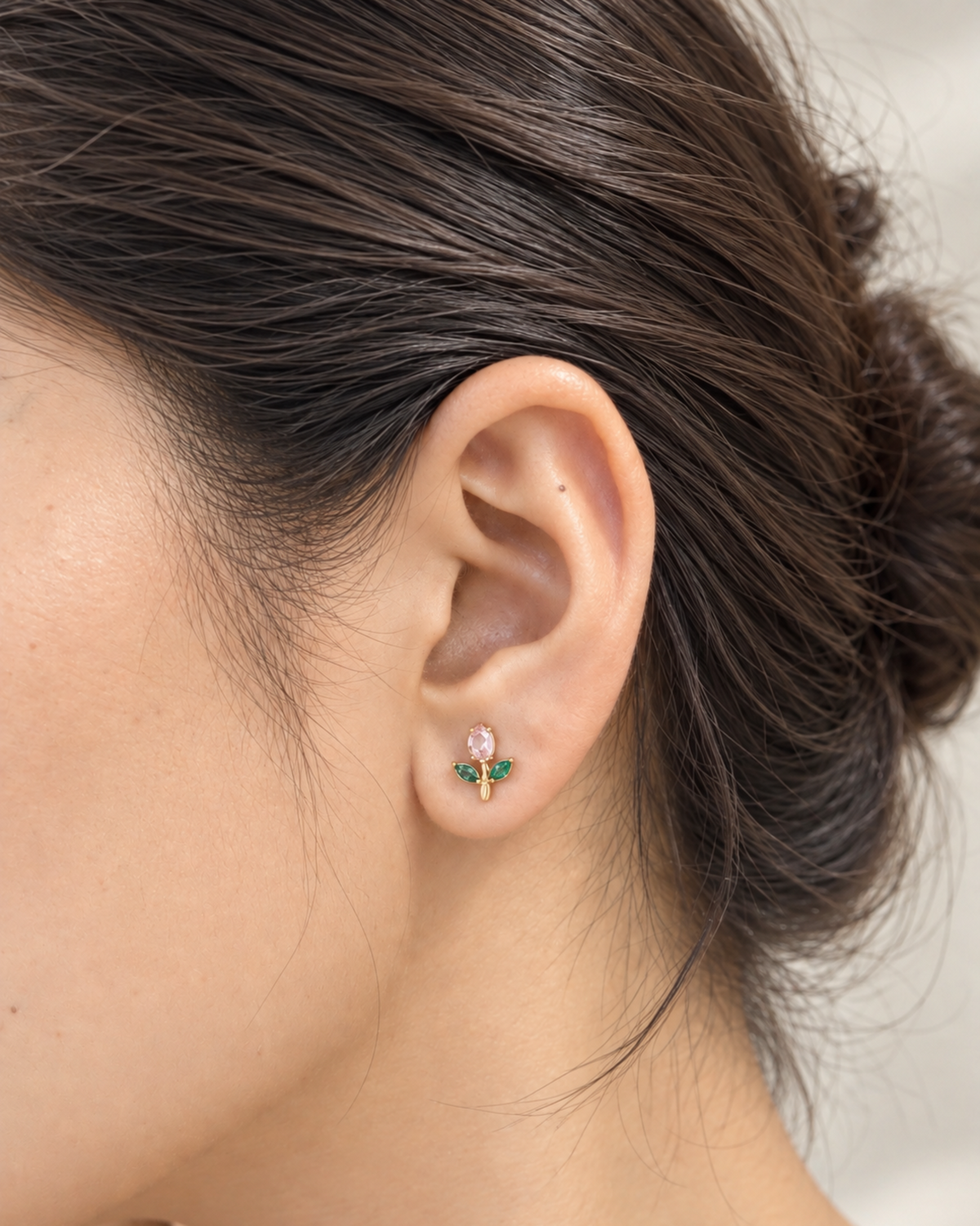 A close-up of a model's ear wearing a small gold stud earring with a pink top and green leaf-like accents.