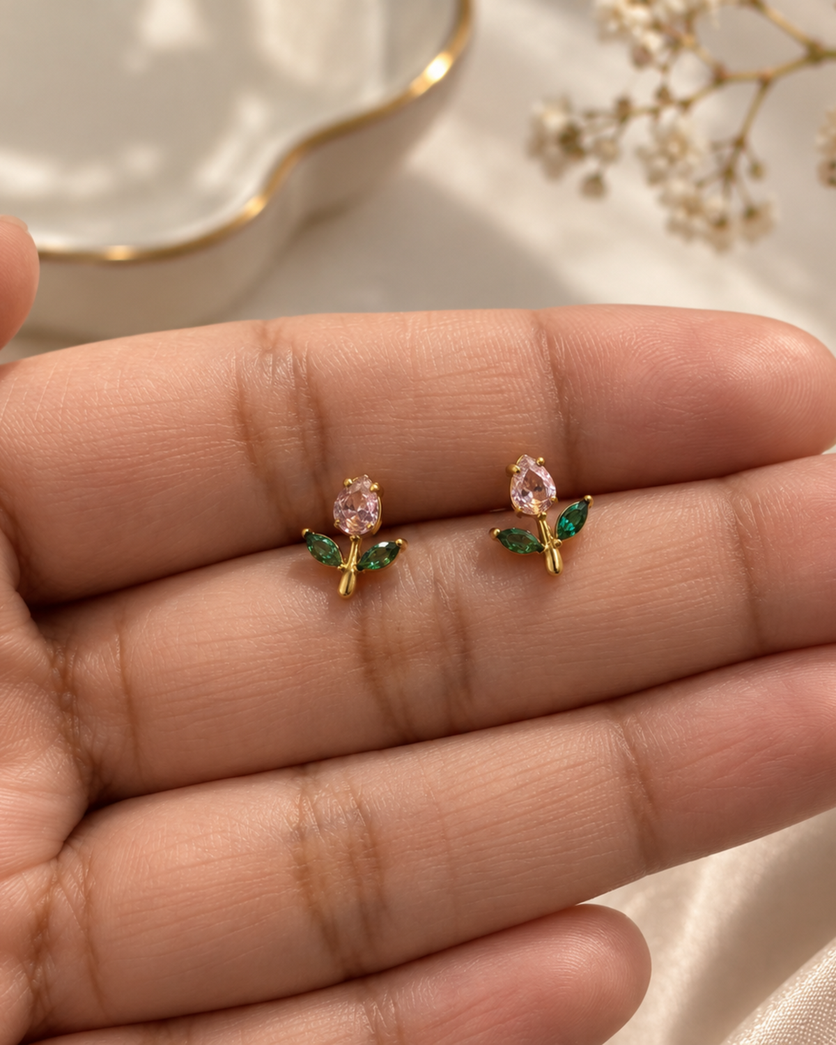 A close-up of a person's hand displaying a pair of small gold flower stud earrings with pink oval center stones and green leaf accents, set against a soft cream satin and dried flower background.