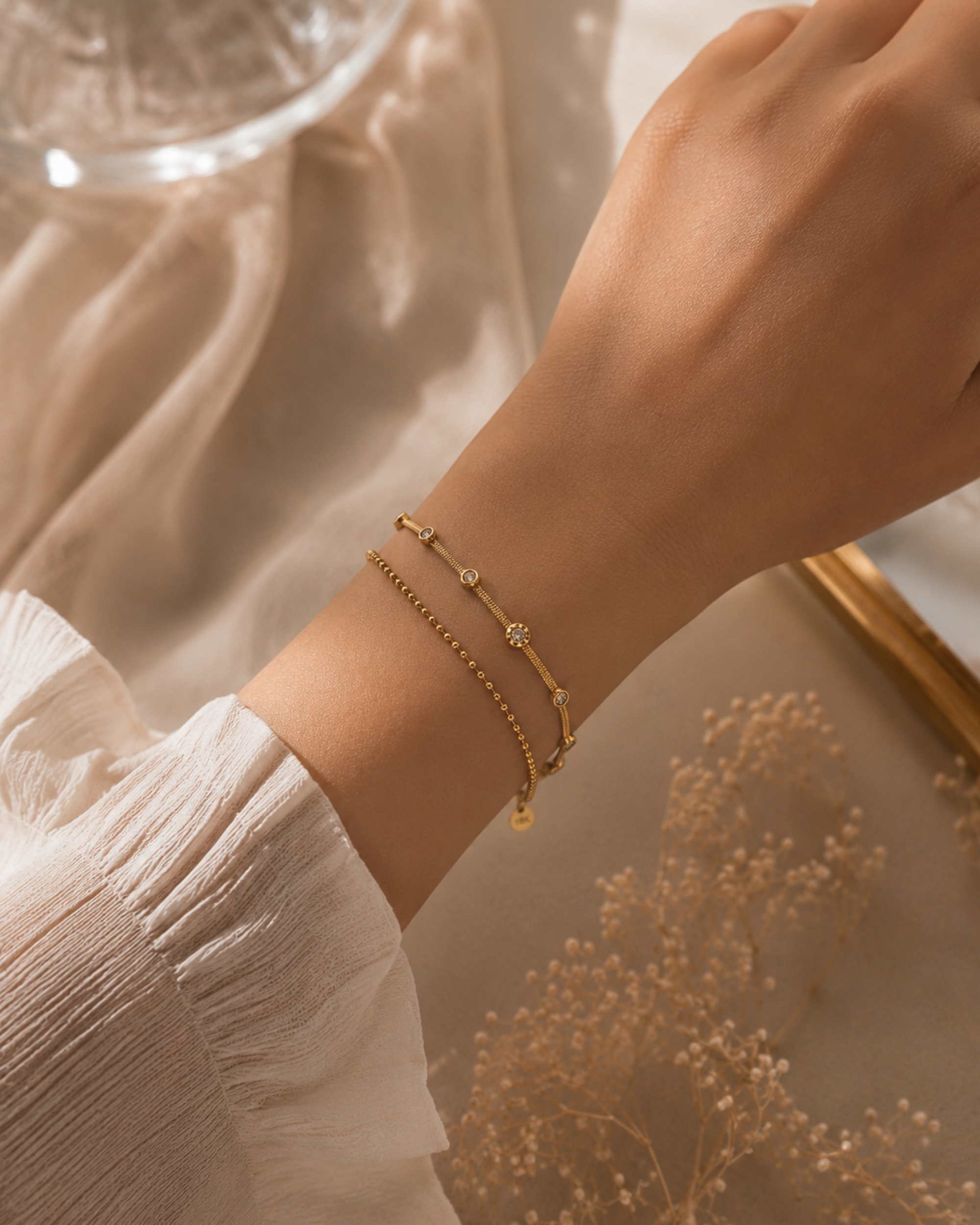 A delicate gold chain bracelet with small bead details worn on a person's wrist, styled with a white ruffled sleeve against a soft beige background with dried flowers.