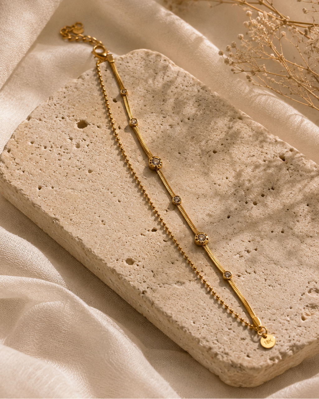 A delicate gold chain anklet with small floral motifs and tiny clear stones, displayed on a textured beige stone slab with soft cream fabric and dried flowers in the background.