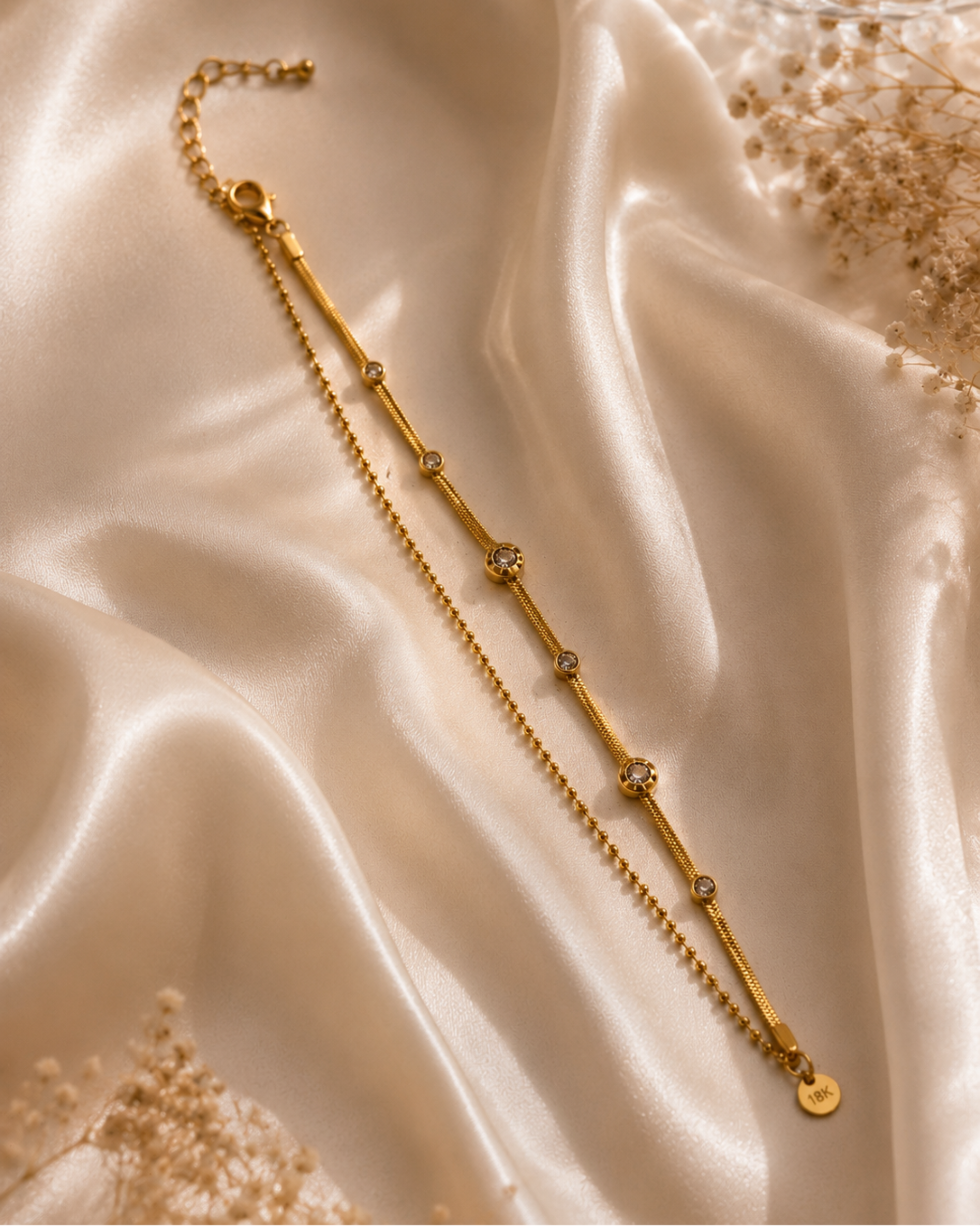 A gold-toned chain anklet with three round clear crystal settings, displayed on draped champagne satin fabric with dried floral accents.