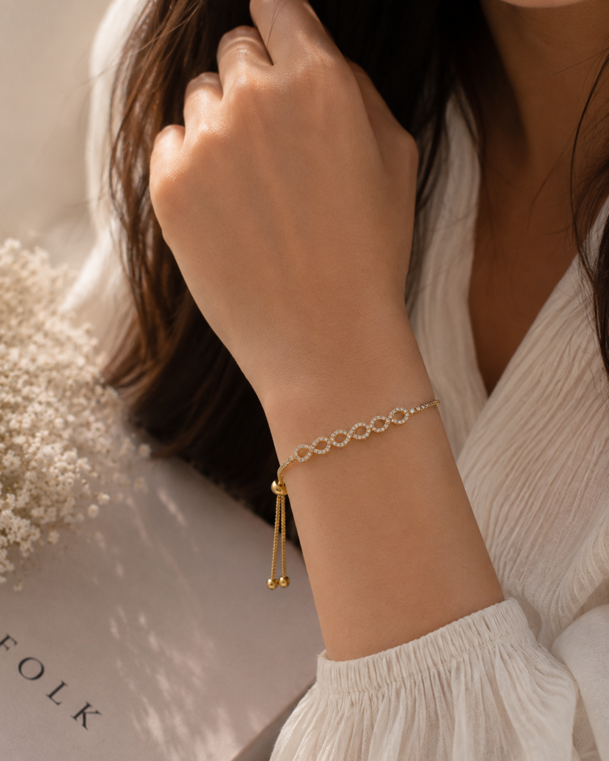 A delicate gold chain bracelet with a safety pin charm and dangling tassels, shown worn on a woman's wrist. The woman is wearing a white blouse, and a soft-focus floral arrangement and a box with the brand name 'FOLK' are visible in the background.