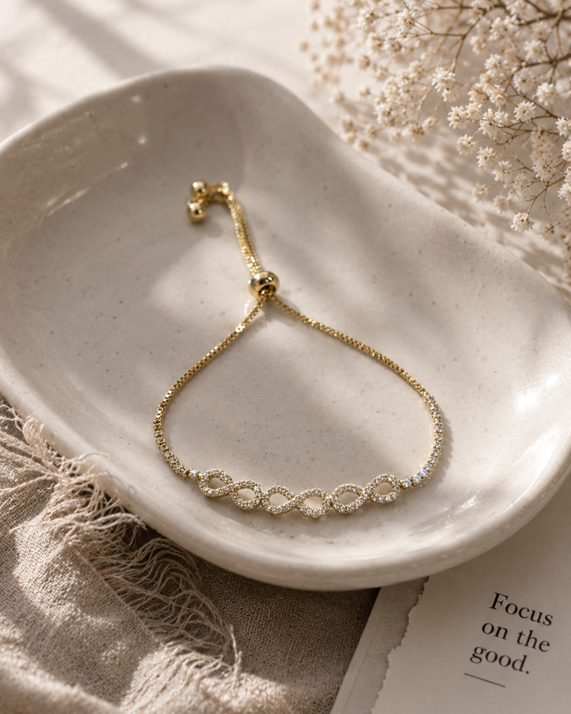 A delicate gold chain bracelet with a row of small clear stones across the center, displayed on a white ceramic dish with neutral fabric and dried flowers around it.